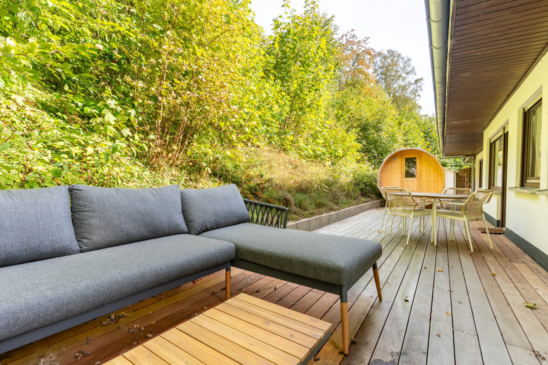 Holzterrasse mit grauem Sofa, Esstisch und runder Sauna, umgeben von viel Grün.