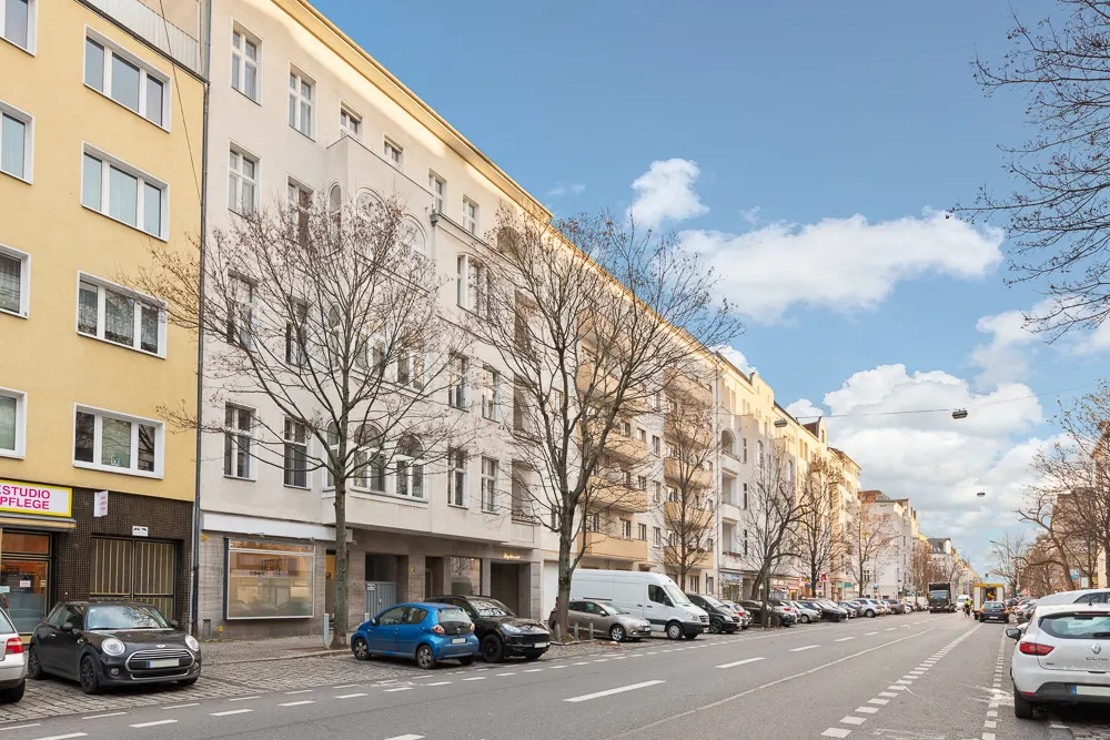 Mehrstöckige Wohnhäuser an einer Straße mit parkenden Autos und kahlen Bäumen.