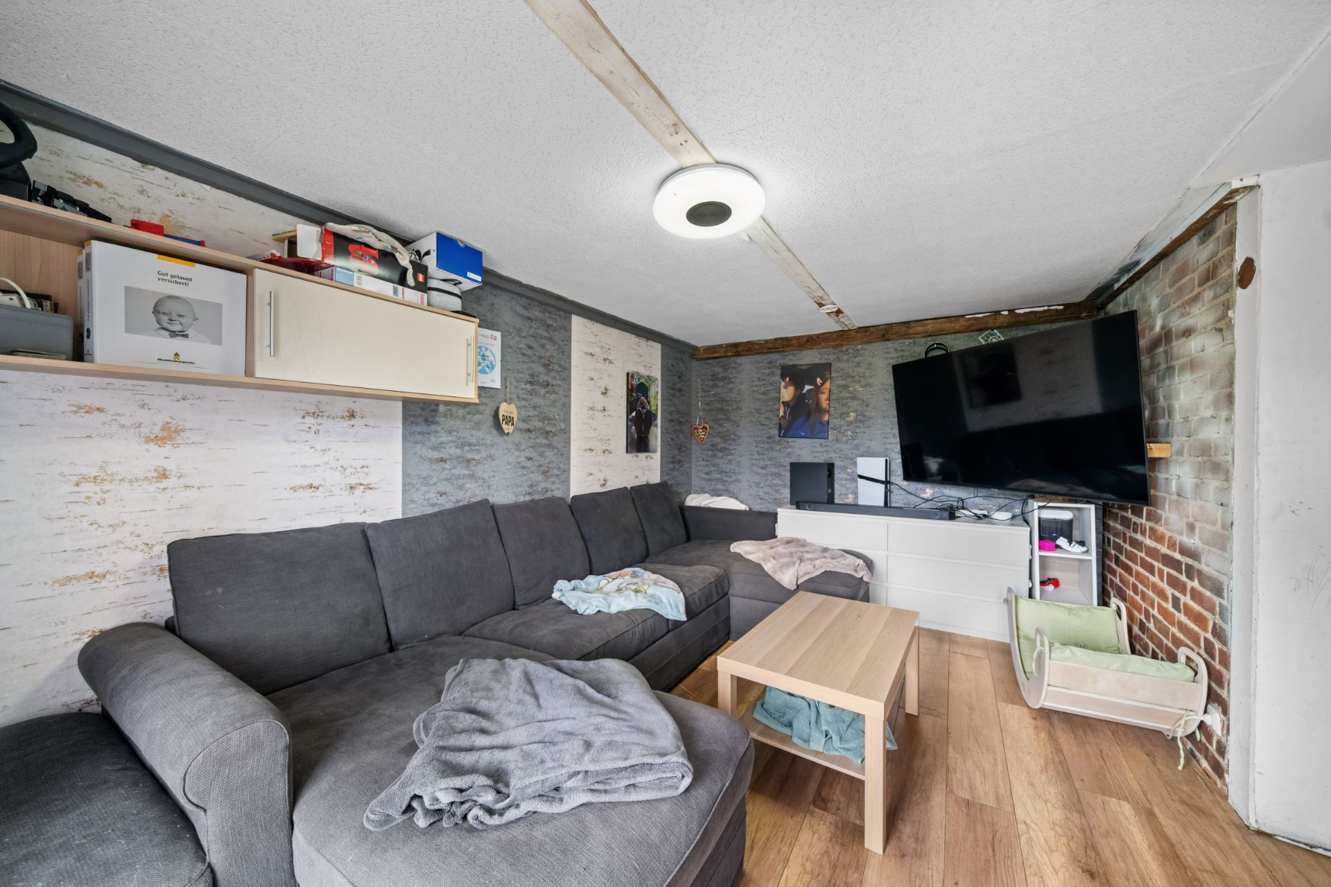 Wohnzimmer mit grauem Ecksofa, Holztisch, TV an der Wand und gemischten Wandverkleidungen.