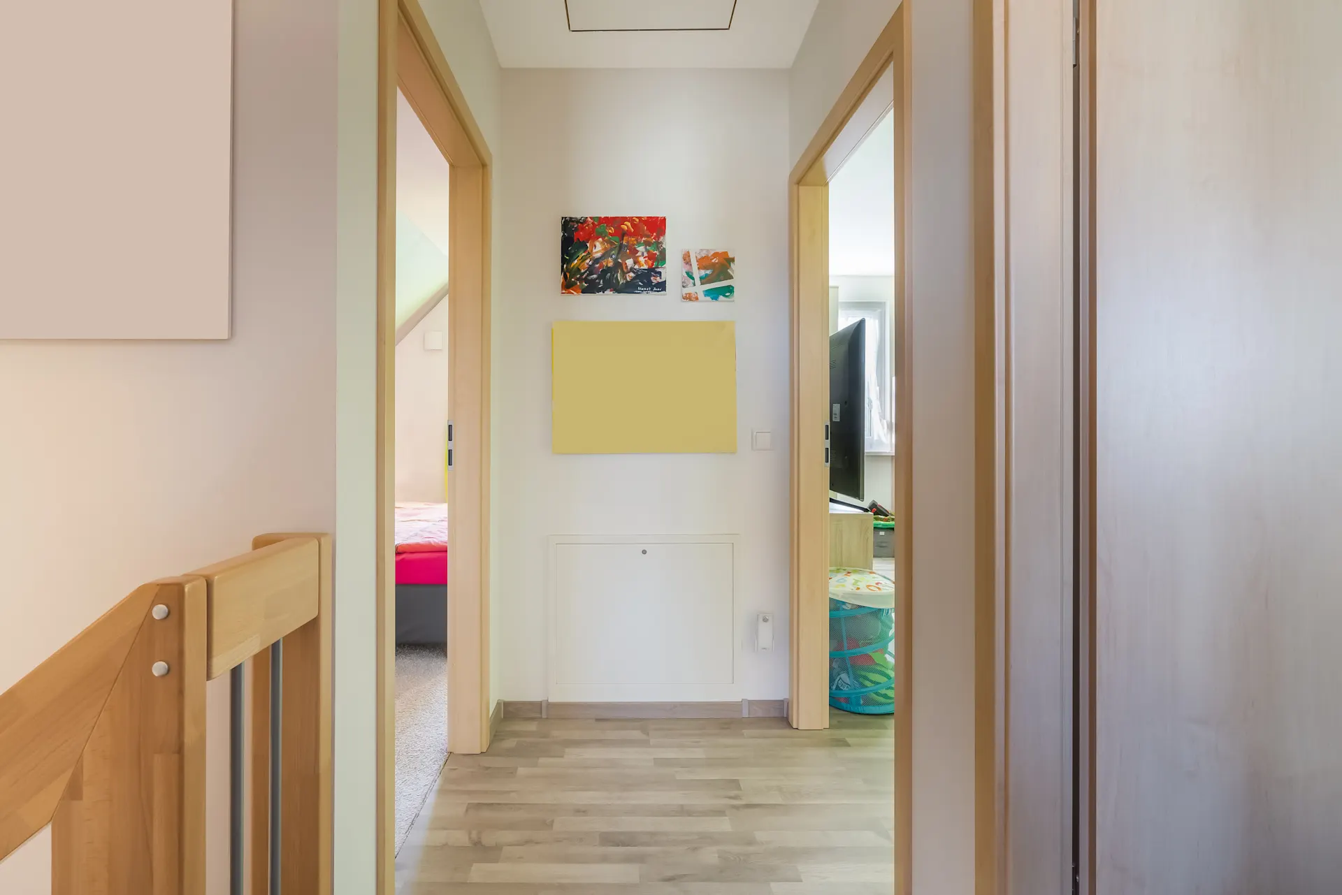 Heller Flur mit Holzboden, zwei offenen Türen und bunten Bildern an der Wand.