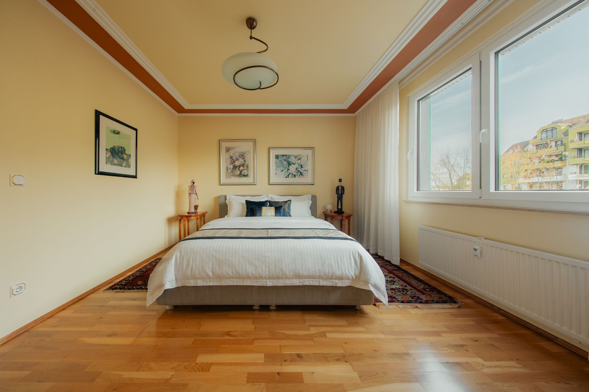 Helles Schlafzimmer mit großem Bett, Holzboden, drei Bildern an der Wand und großen Fenstern.