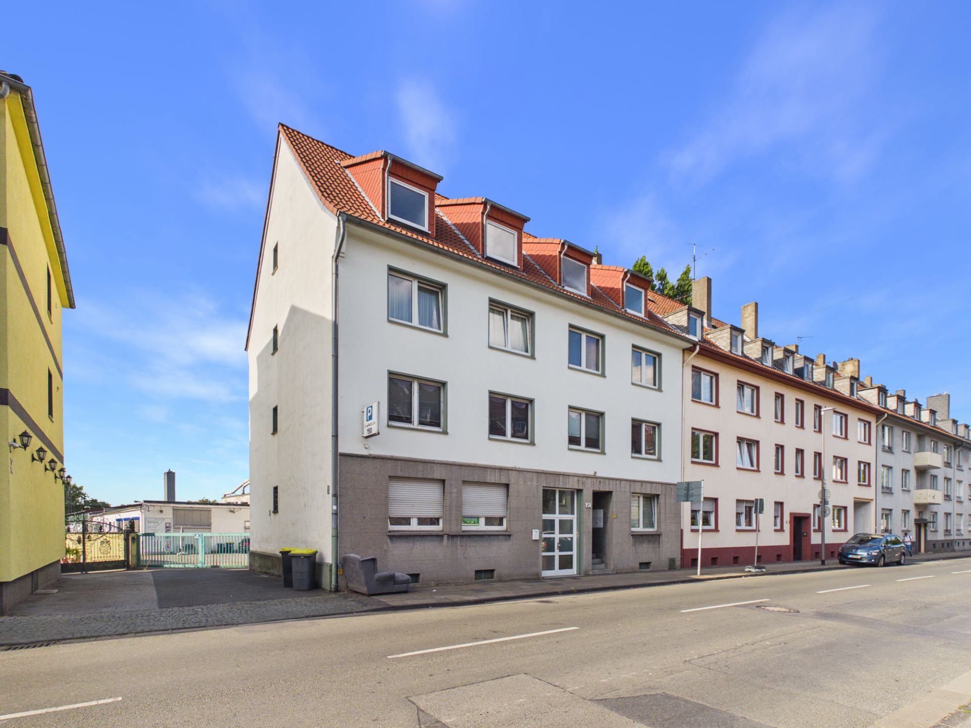 Mehrstöckiges Wohnhaus mit rotem Dach an einer Straße, mehrere Fenster und Eingänge sichtbar.