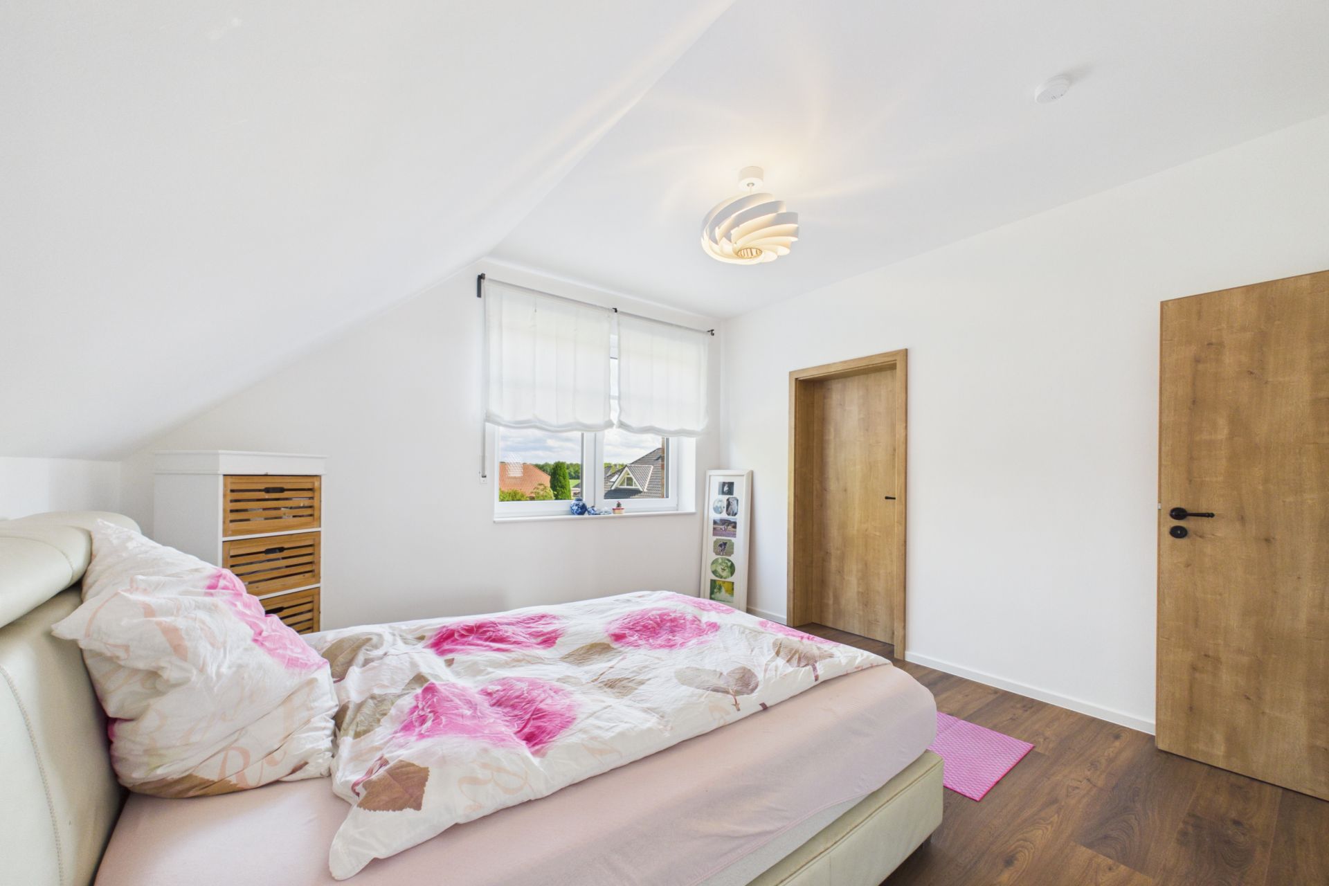 Helles Schlafzimmer mit Bett, weißer Bettwäsche, Holzkommode und Fenster mit Rollos.