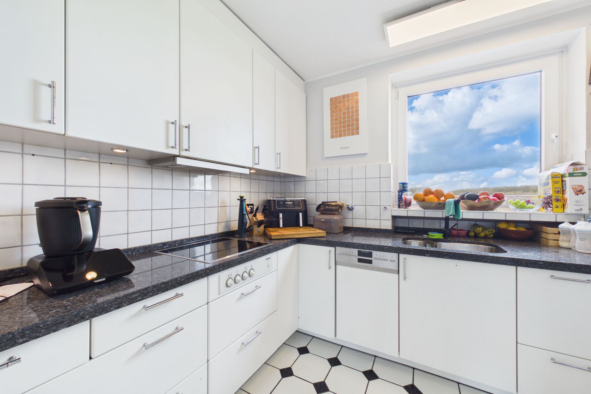 Helle Küche mit weißen Schränken, Elektrogeräten und einem Fenster mit Ausblick.