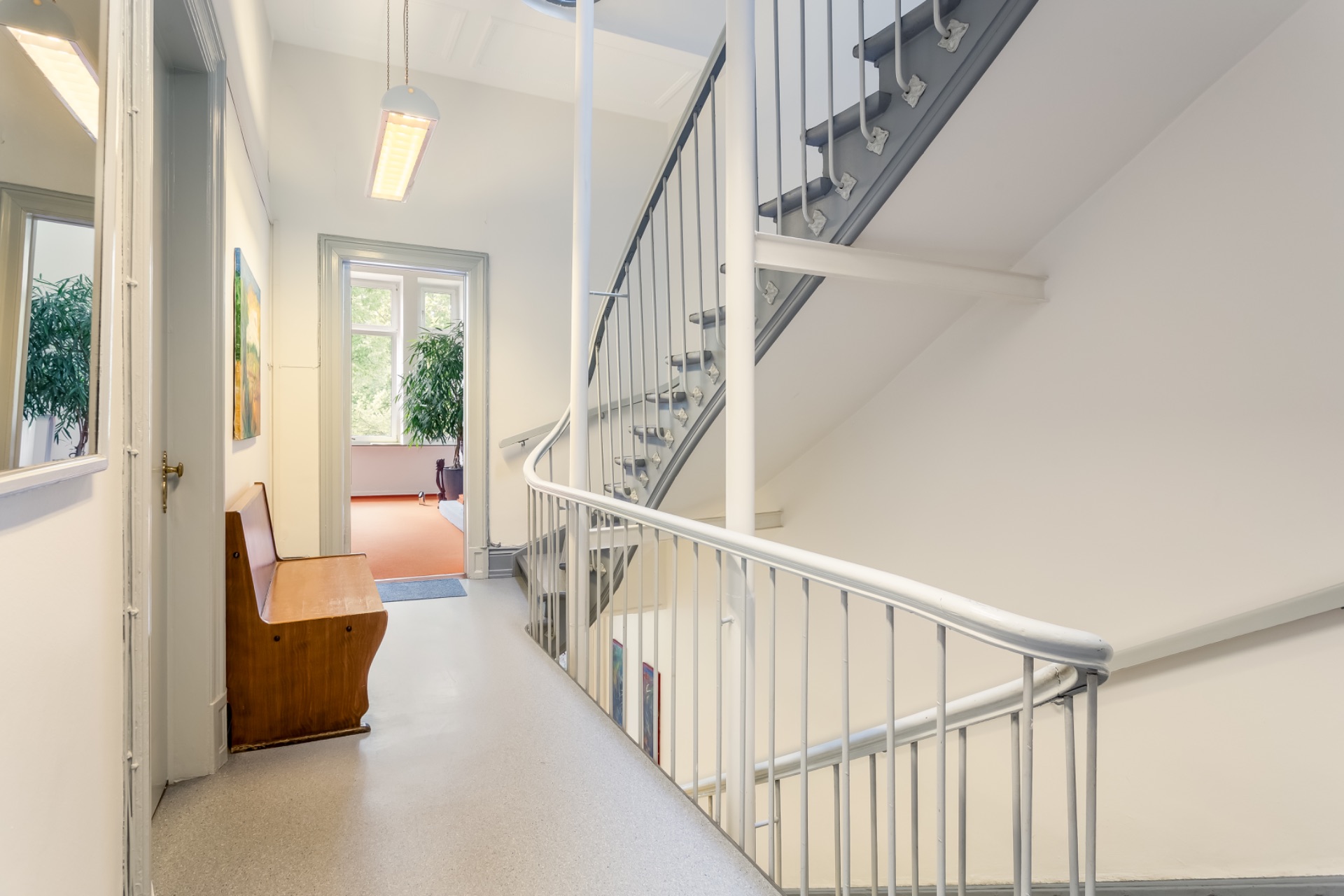 Heller Flur mit Holzbank, Treppe und Zugang zu einem Raum mit großen Fenstern und Pflanze.