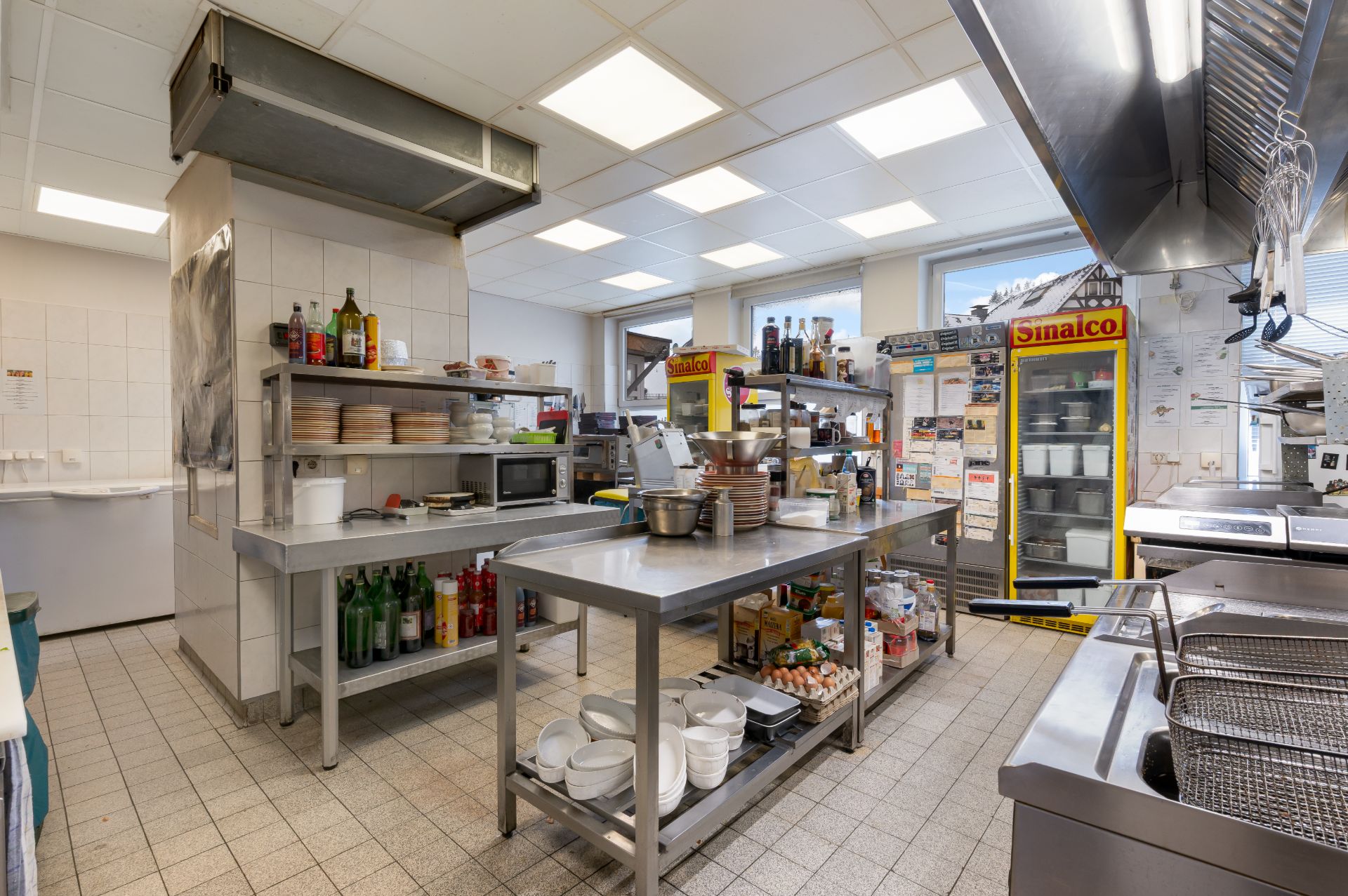 Große Küche mit Edelstahltischen, Regalen, Kühlschrank und Kochutensilien.