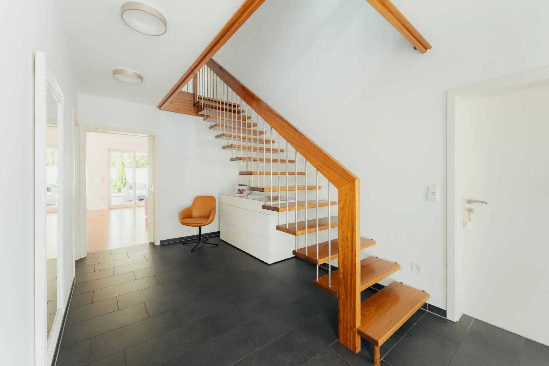 Heller Flur mit Holztreppe, dunklem Boden, weißer Kommode und orangefarbenem Stuhl.