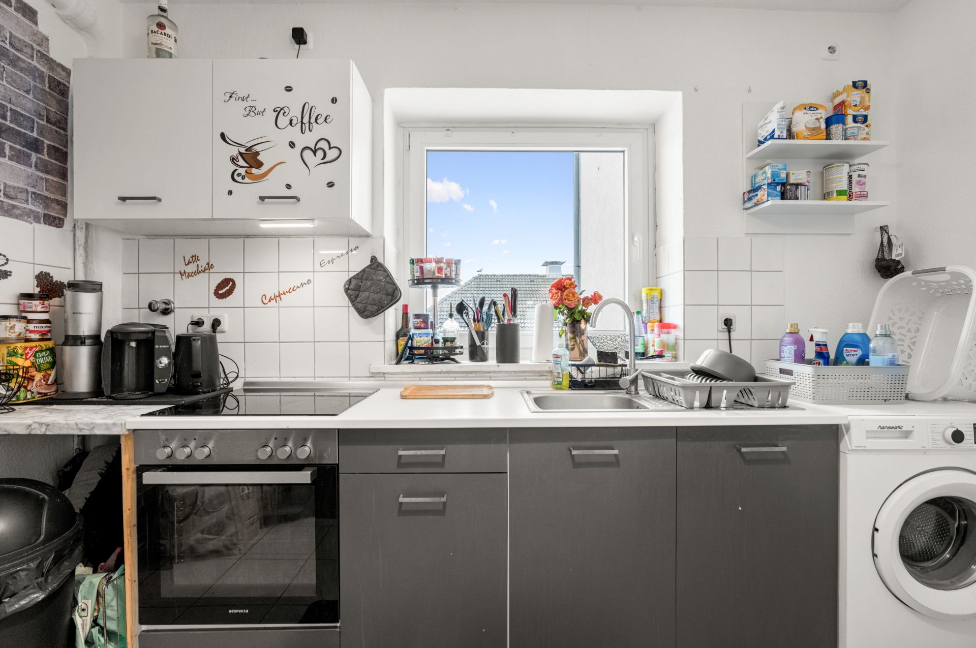 Helle Küche mit Herd, Spüle, Waschmaschine, Regalen und Küchenutensilien vor einem Fenster.