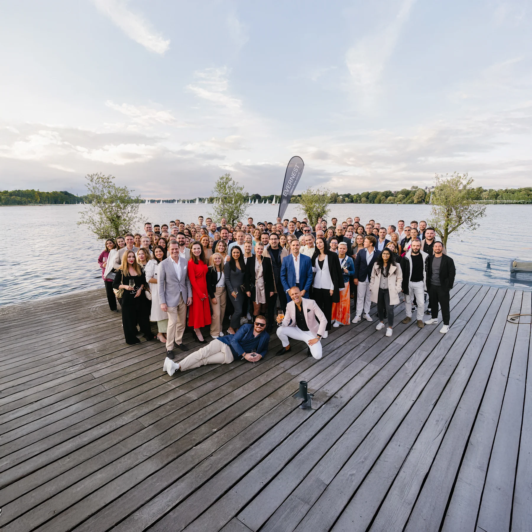 Große Gruppe von Menschen steht auf einem Holzsteg an einem See mit Bäumen im Hintergrund.