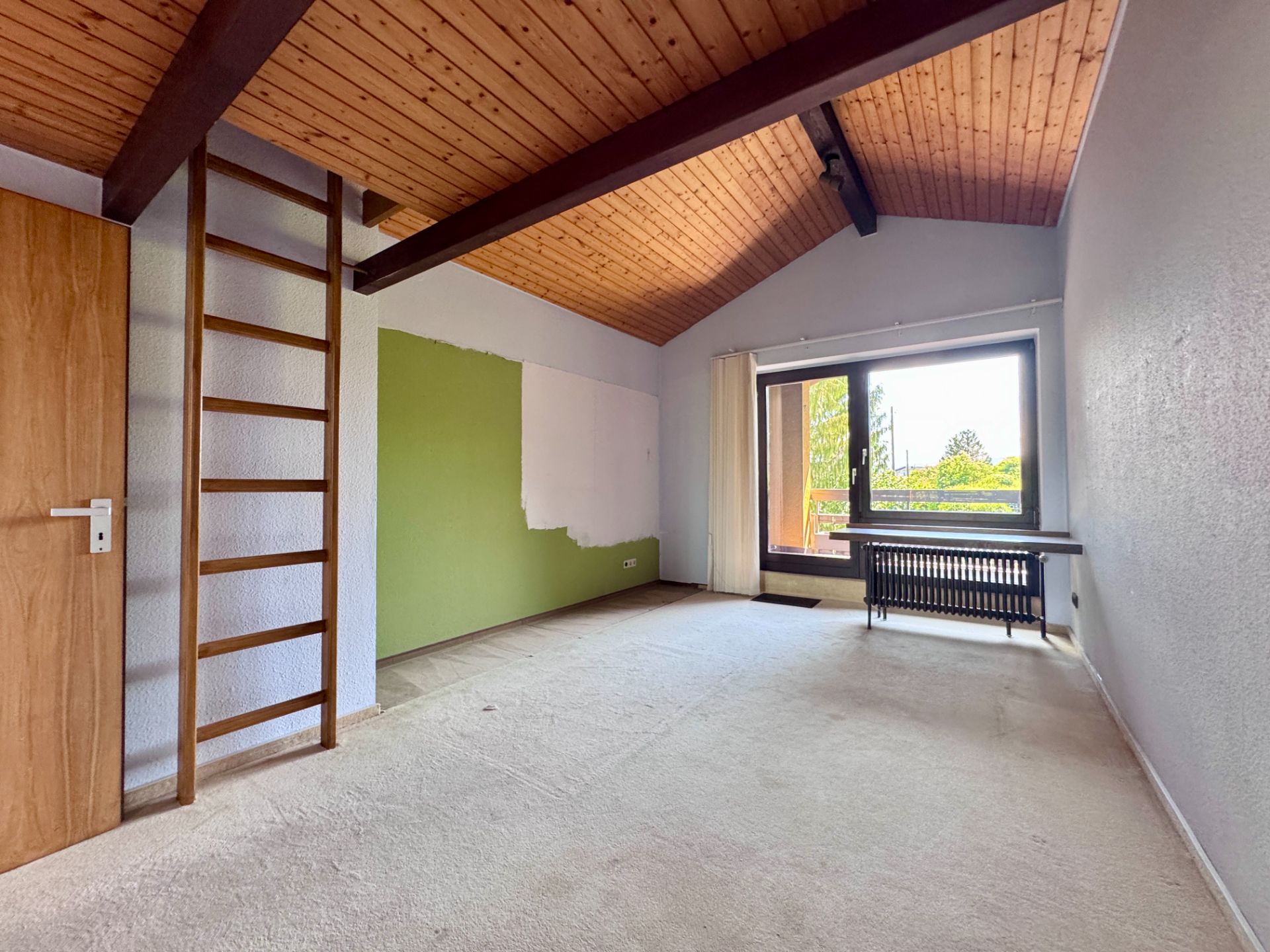 Leerer Raum mit Teppichboden, Holzdecke, Leiter, großem Fenster und Heizung.
