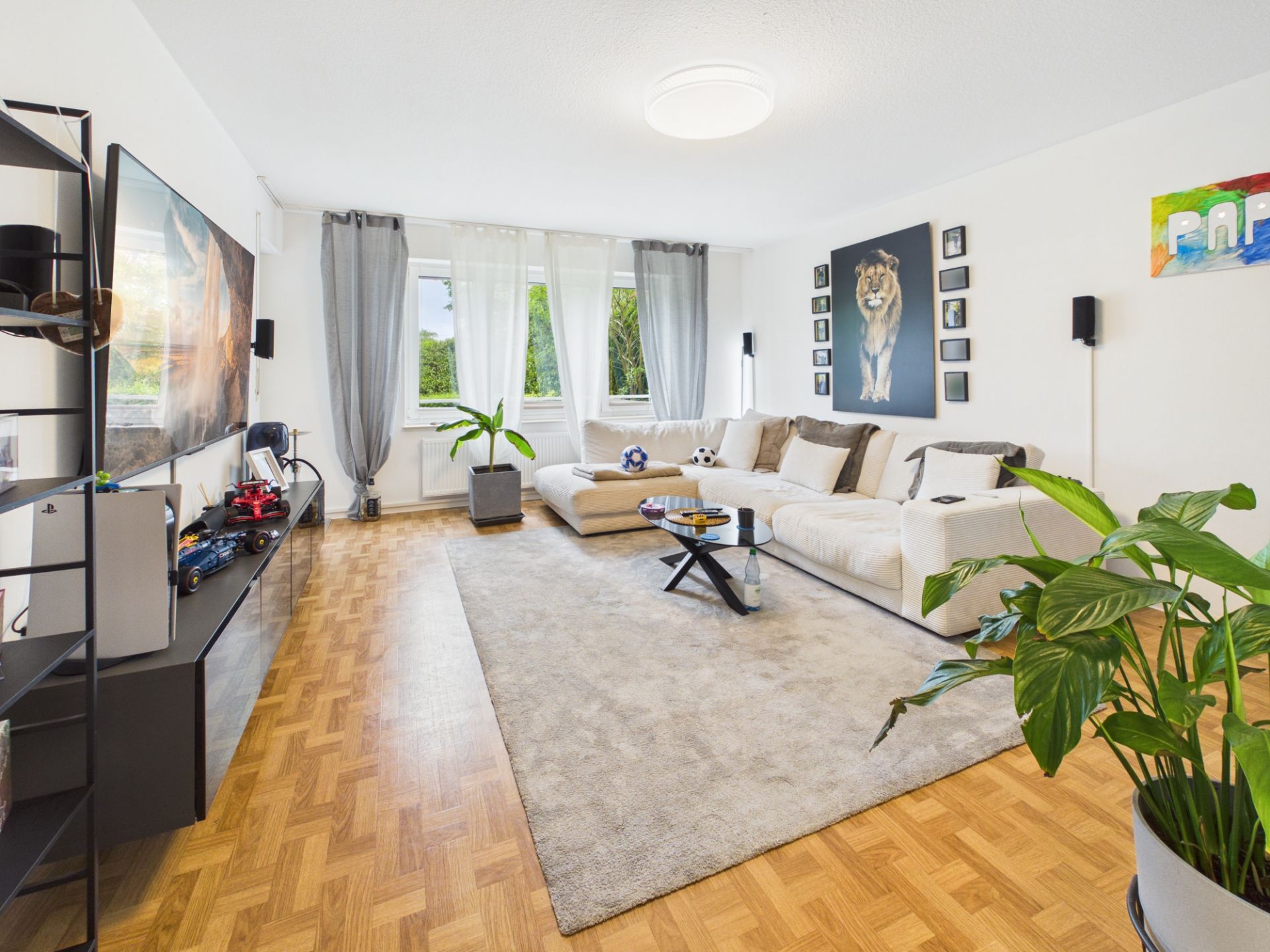 Modernes Wohnzimmer mit großem Sofa, Teppich, Fernseher und Pflanzen, helle Fenster im Hintergrund.