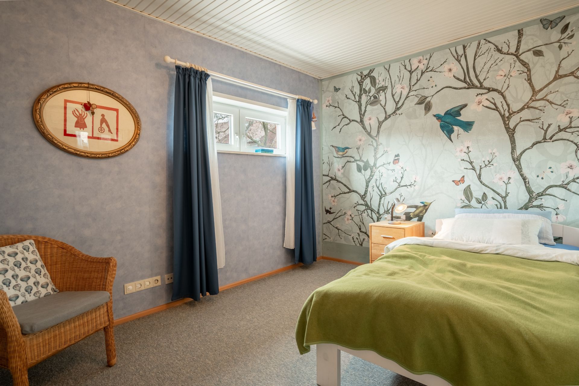 Schlafzimmer mit grünem Bett, Wandgemälde mit Vögeln, Fenster, Rattanstuhl und Nachttisch.