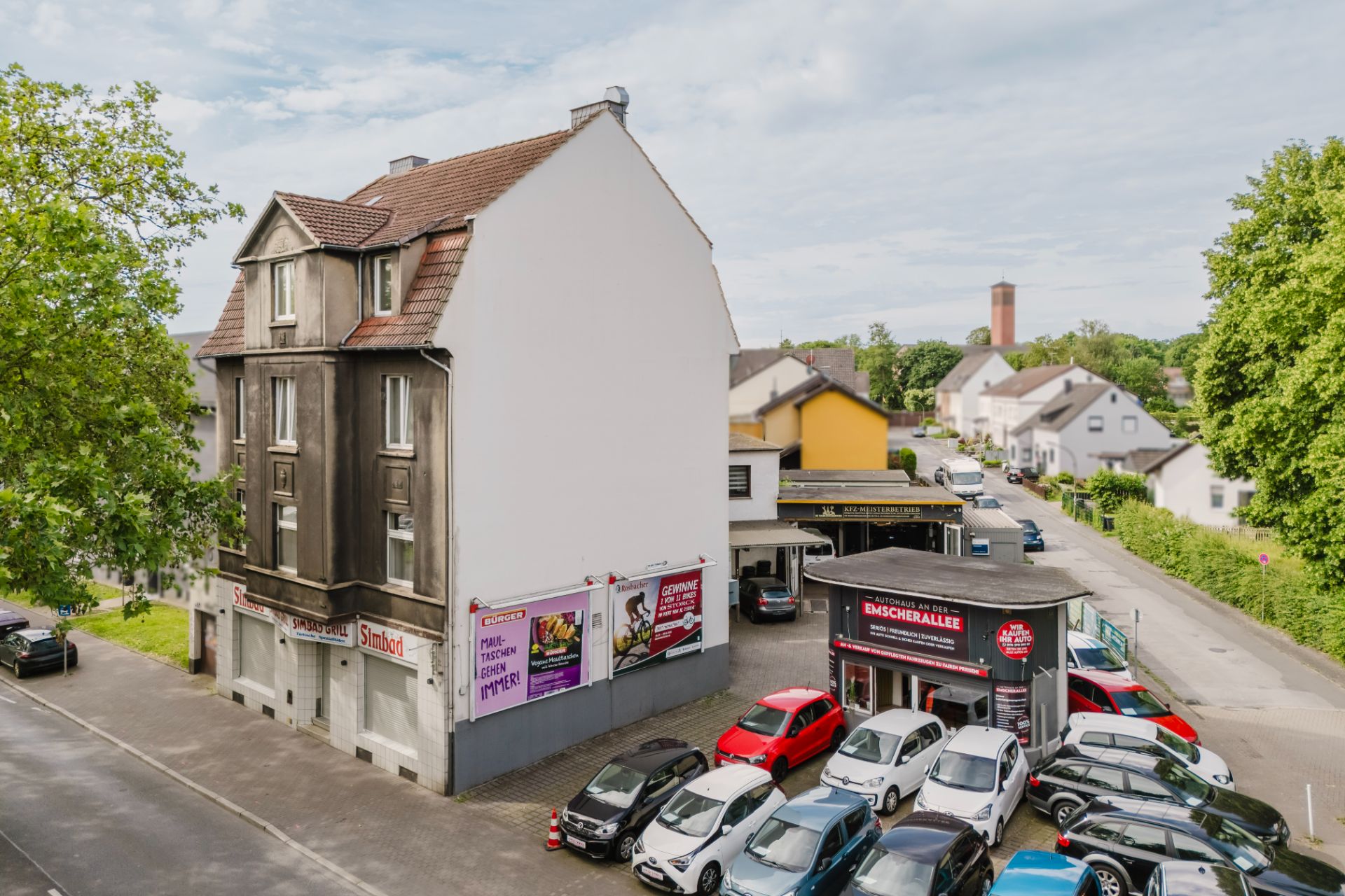 Mehrfamilienhaus an einer Straße mit mehreren geparkten Autos und kleinem Laden im Vordergrund.