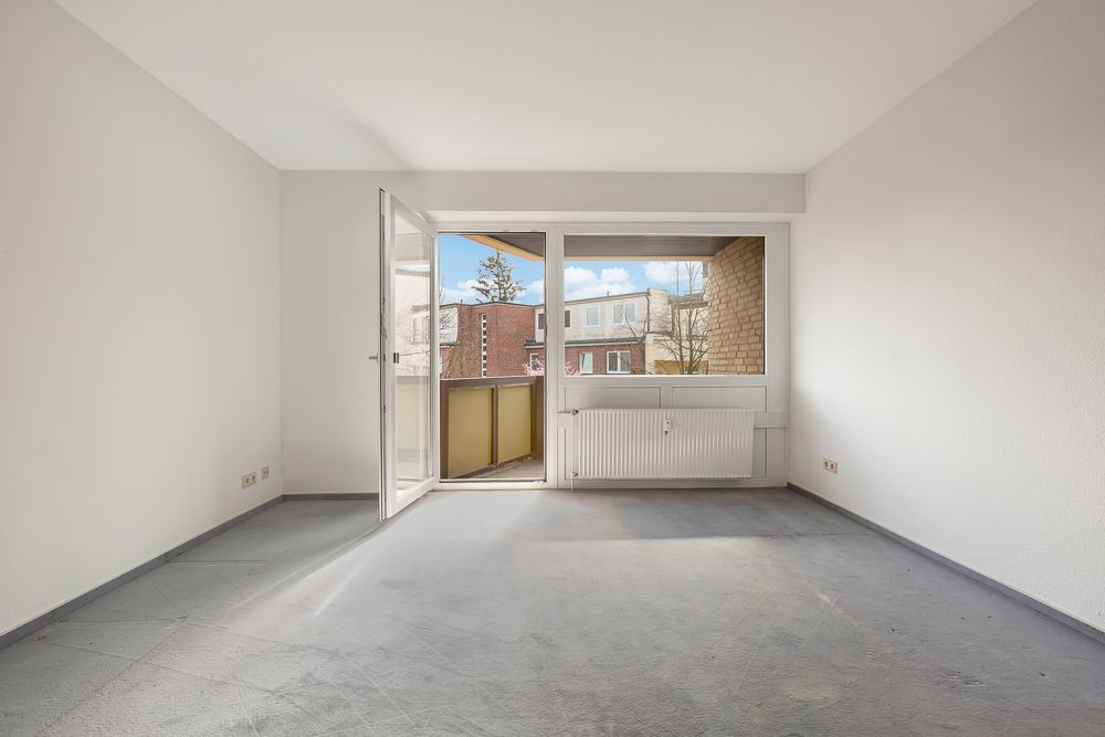 Leeres Zimmer mit grauem Boden, weißen Wänden und offenem Zugang zu einem kleinen Balkon.