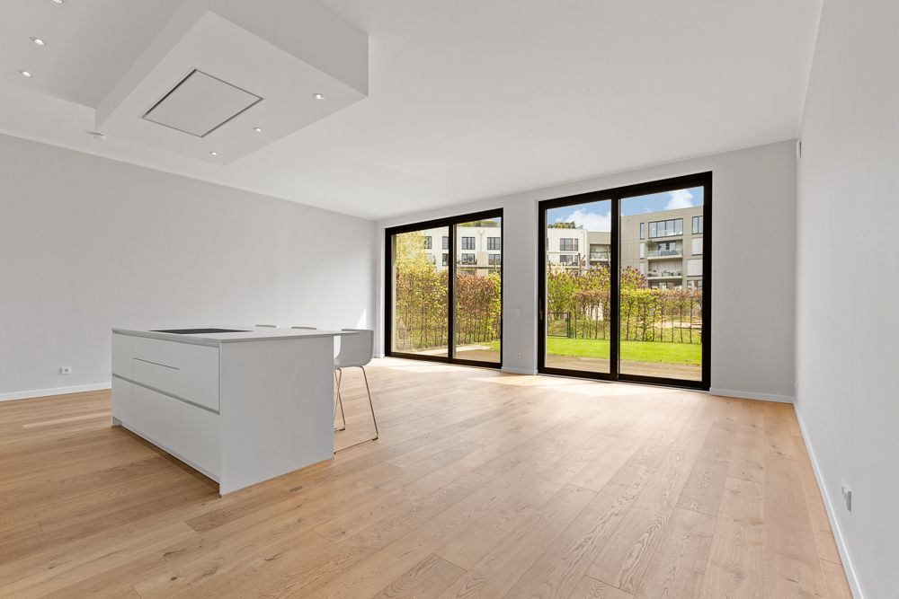 Helles, leeres Wohnzimmer mit offener Küche, Holzboden und großen Fenstern zum Garten.