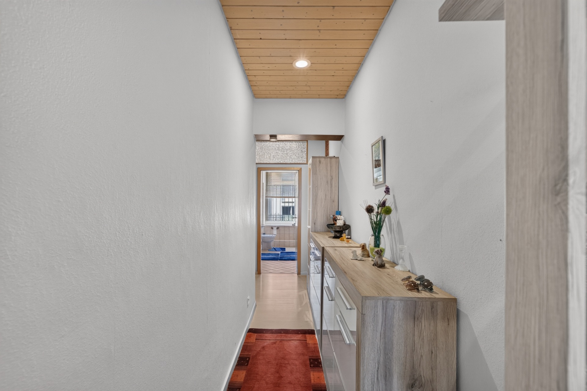 Schmaler Flur mit Holzdecke, Kommode, rotem Teppich und Blick in ein weiteres Zimmer.