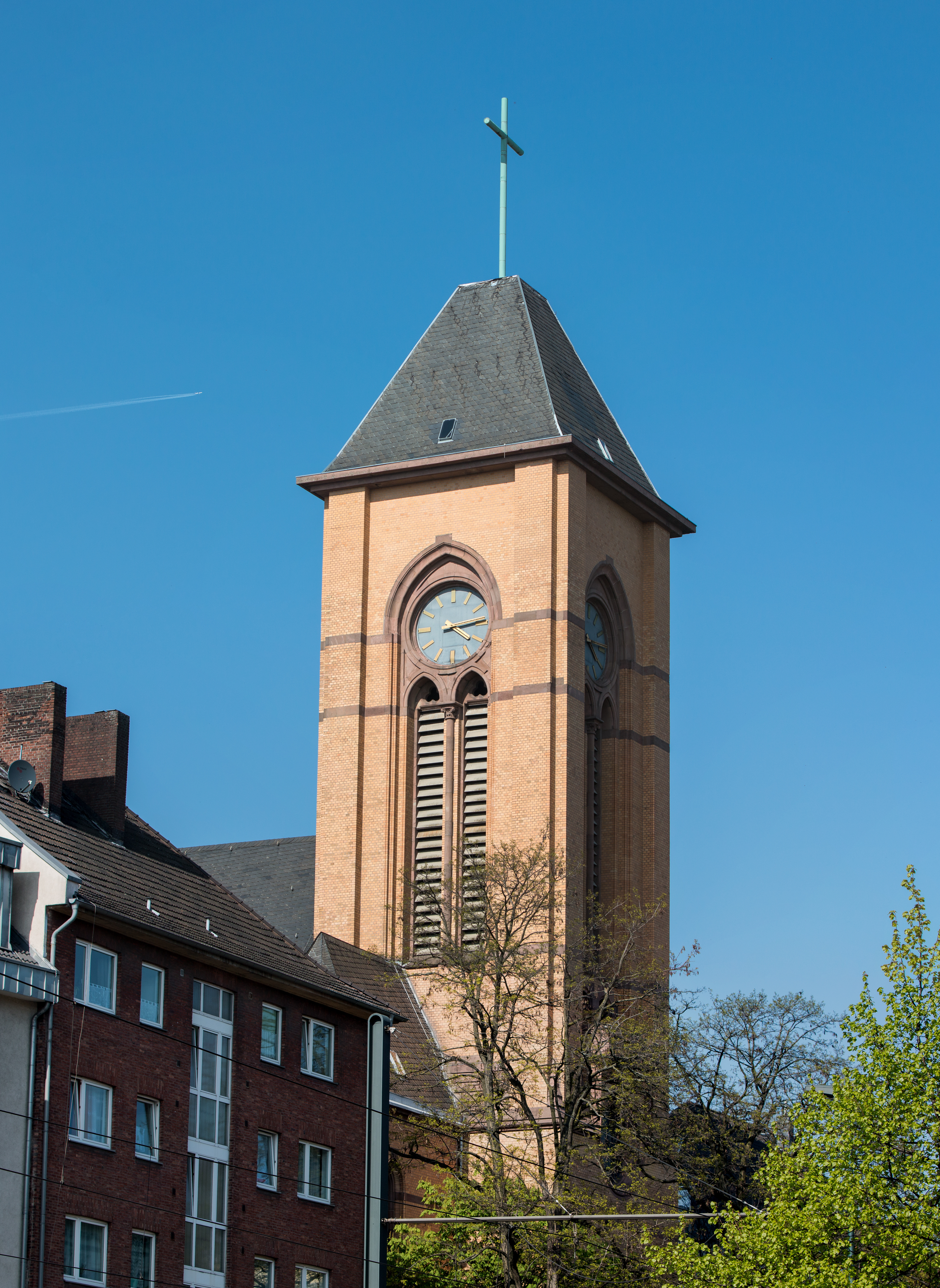 Platz mit Rundturm, Bäumen und mehrstöckigem Wohngebäude bei blauem Himmel.