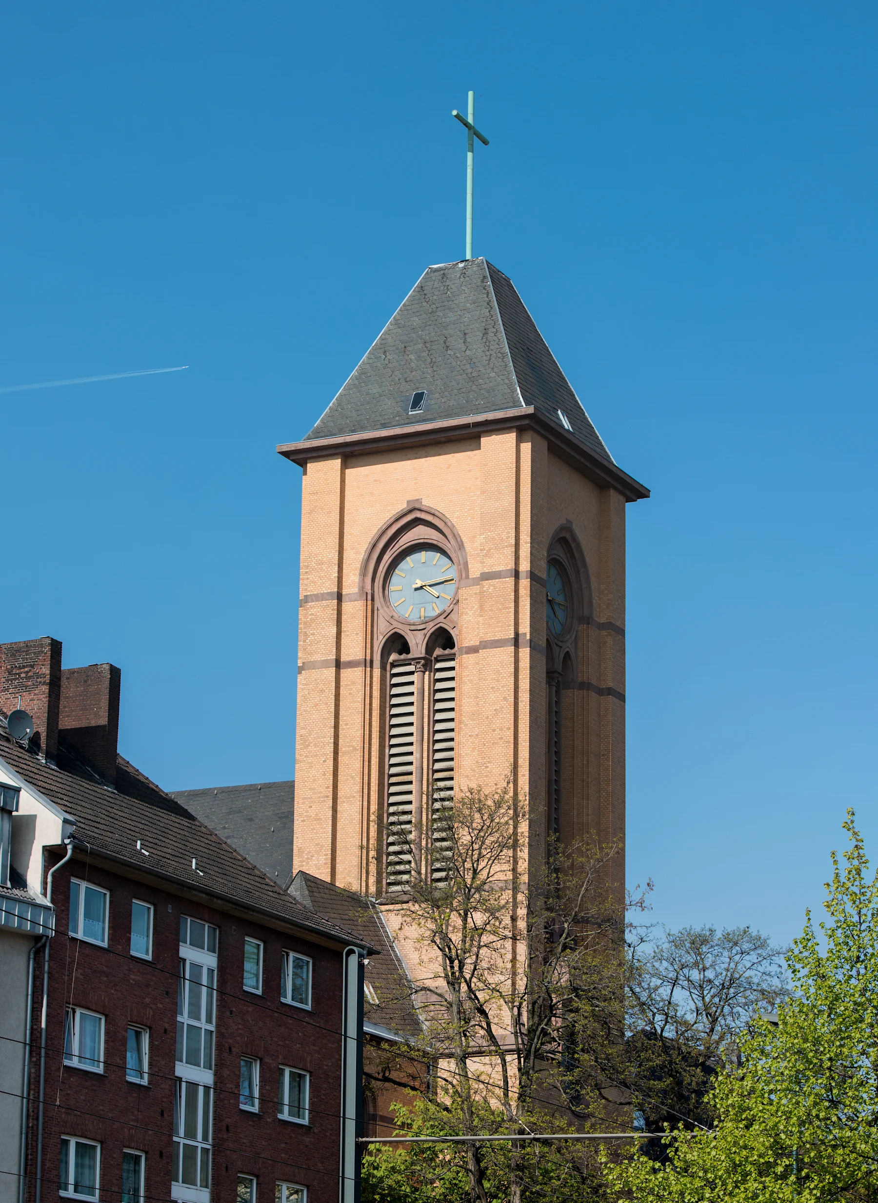 Platz mit Rundturm, Bäumen und mehrstöckigem Wohngebäude bei blauem Himmel.