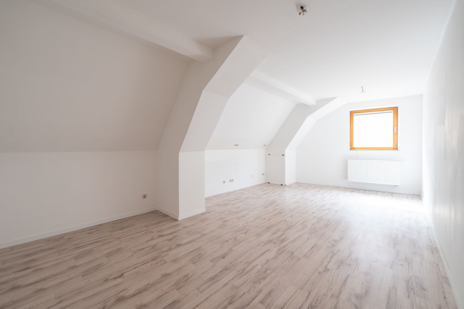 Heller leerer Raum mit schrägen Wänden, Laminatboden und einem Fenster mit Heizkörper.
