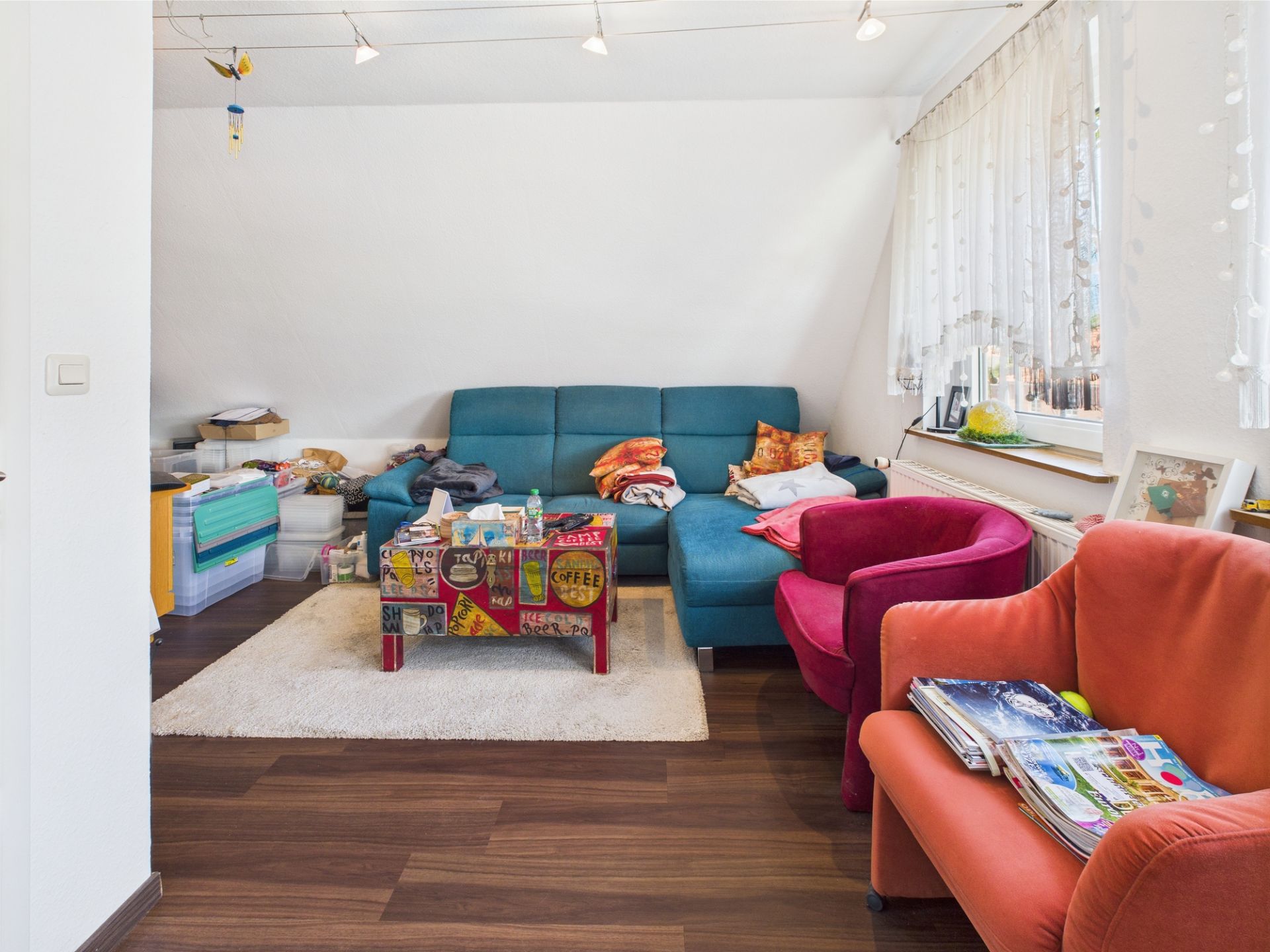Wohnzimmer mit blauem Sofa, buntem Couchtisch und zwei Sesseln, Fenster mit Gardine rechts.