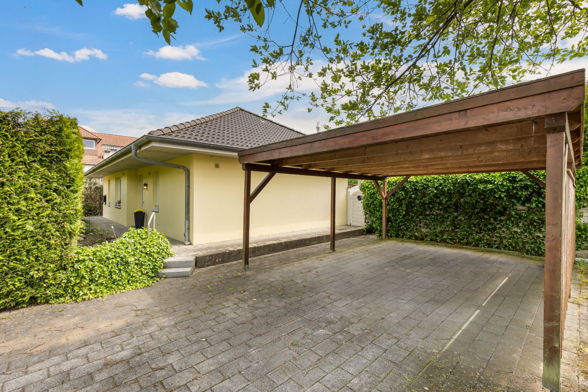 Gelbes Einfamilienhaus mit dunklem Dach und einem Carport aus Holz auf gepflastertem Hof.