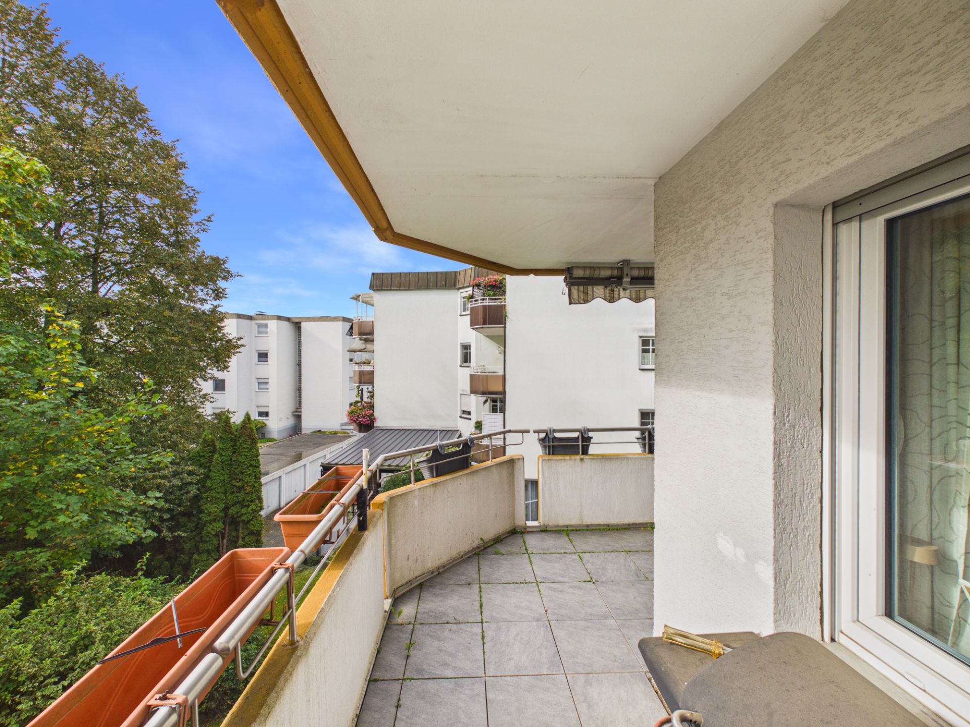 Balkon mit Fliesenboden, Blumenkästen und Blick auf benachbarte Wohngebäude und Bäume.