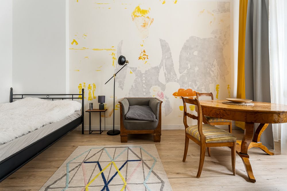 Helles Zimmer mit Einzelbett, Sessel, Holztisch, zwei Stühlen und gemusterter Wand.