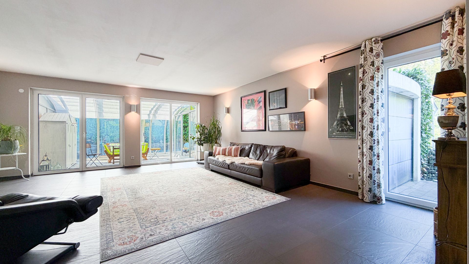 Helles Wohnzimmer mit großem Teppich, braunem Sofa und Zugang zur Terrasse durch Glastüren.