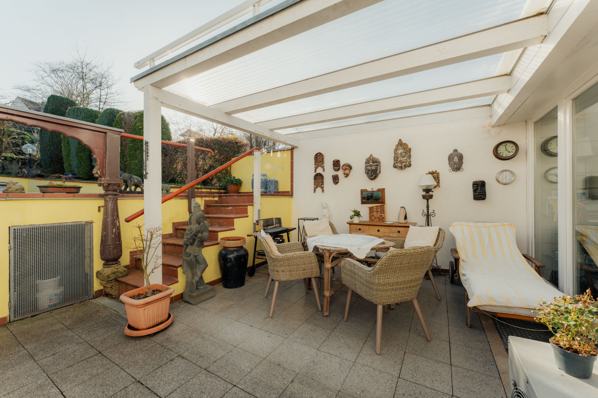 Überdachte Terrasse mit Esstisch, Stühlen und Liege, Wanddekorationen und Pflanzen.