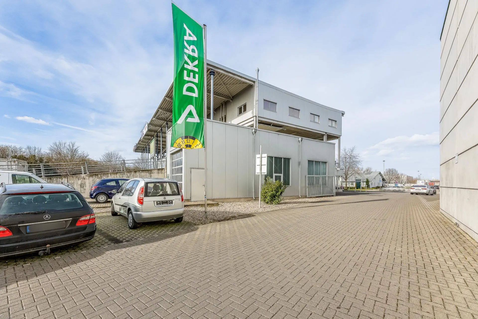 Außenansicht eines modernen Gebäudes mit DEKRA-Fahne und geparkten Autos auf einem gepflasterten Hof.