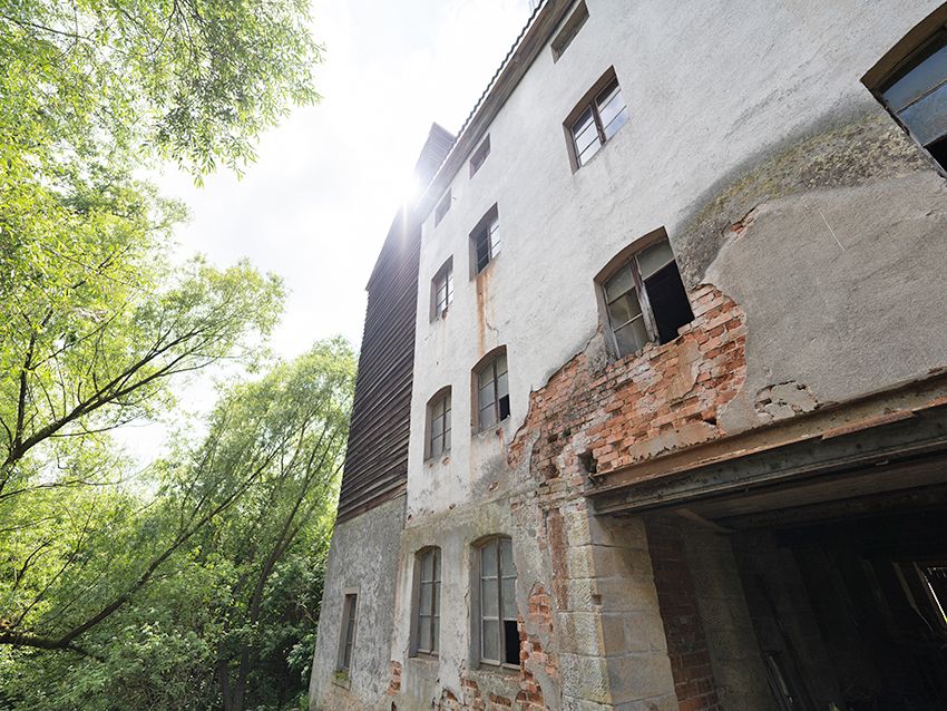 Altes, mehrstöckiges Wohnhaus mit beschädigter Fassade und teils geöffneten Fenstern neben Bäumen.