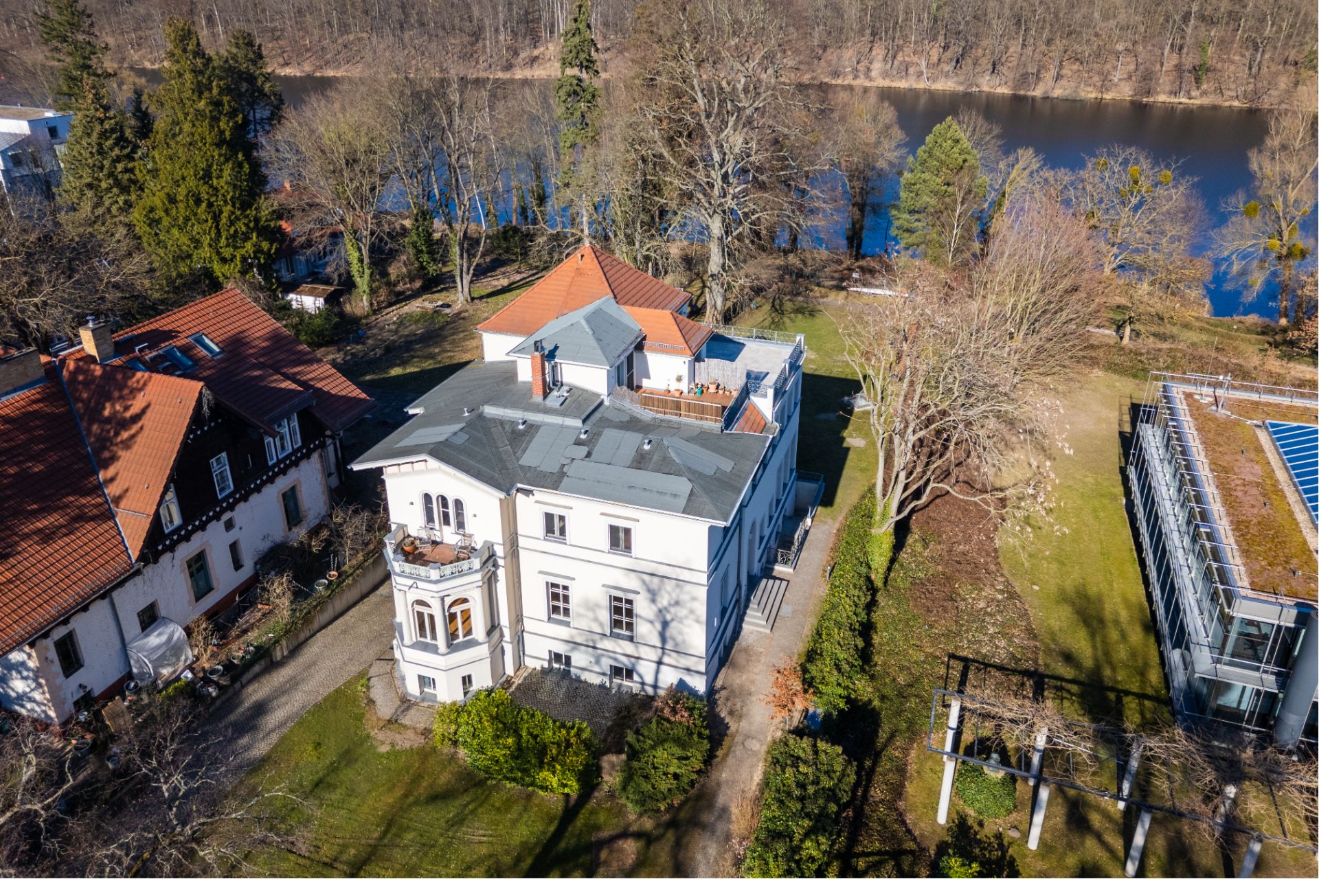 Luftaufnahme eines großen Wohnhauses mit Garten, Nachbargebäuden und einem Fluss im Hintergrund.