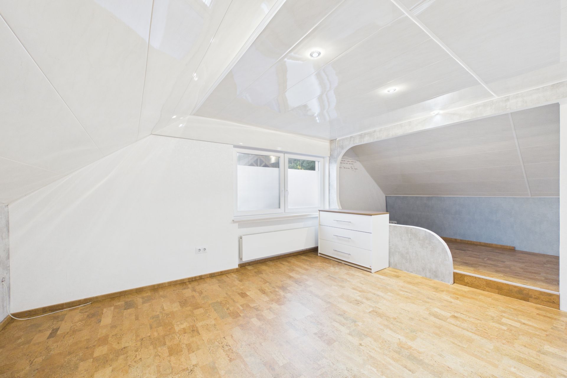 Heller Raum mit schrägen Wänden, Holzfußboden, Fenster und weißer Kommode.
