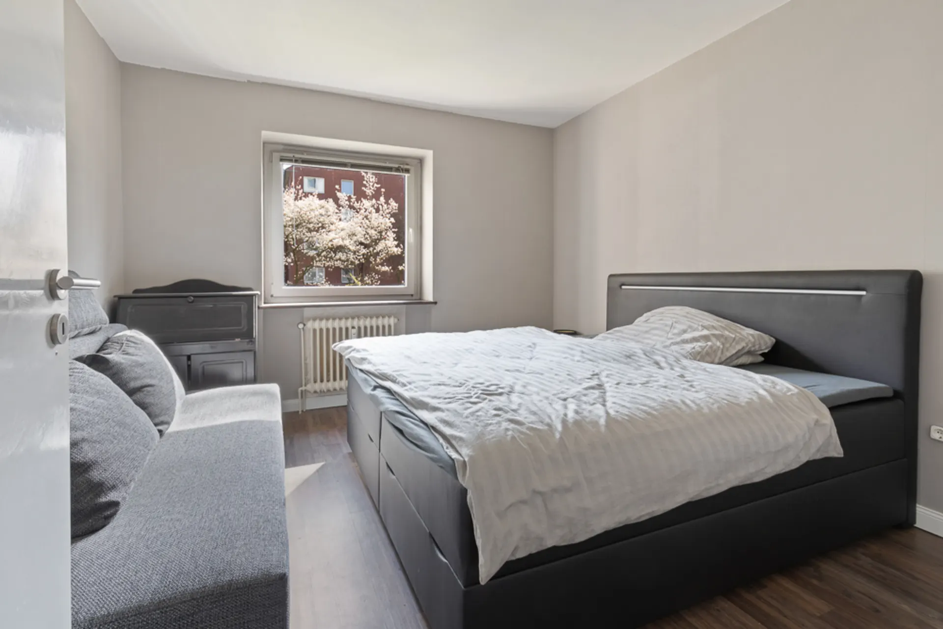 Schlafzimmer mit Doppelbett, grauem Sofa und Fenster mit Blick auf blühenden Baum.