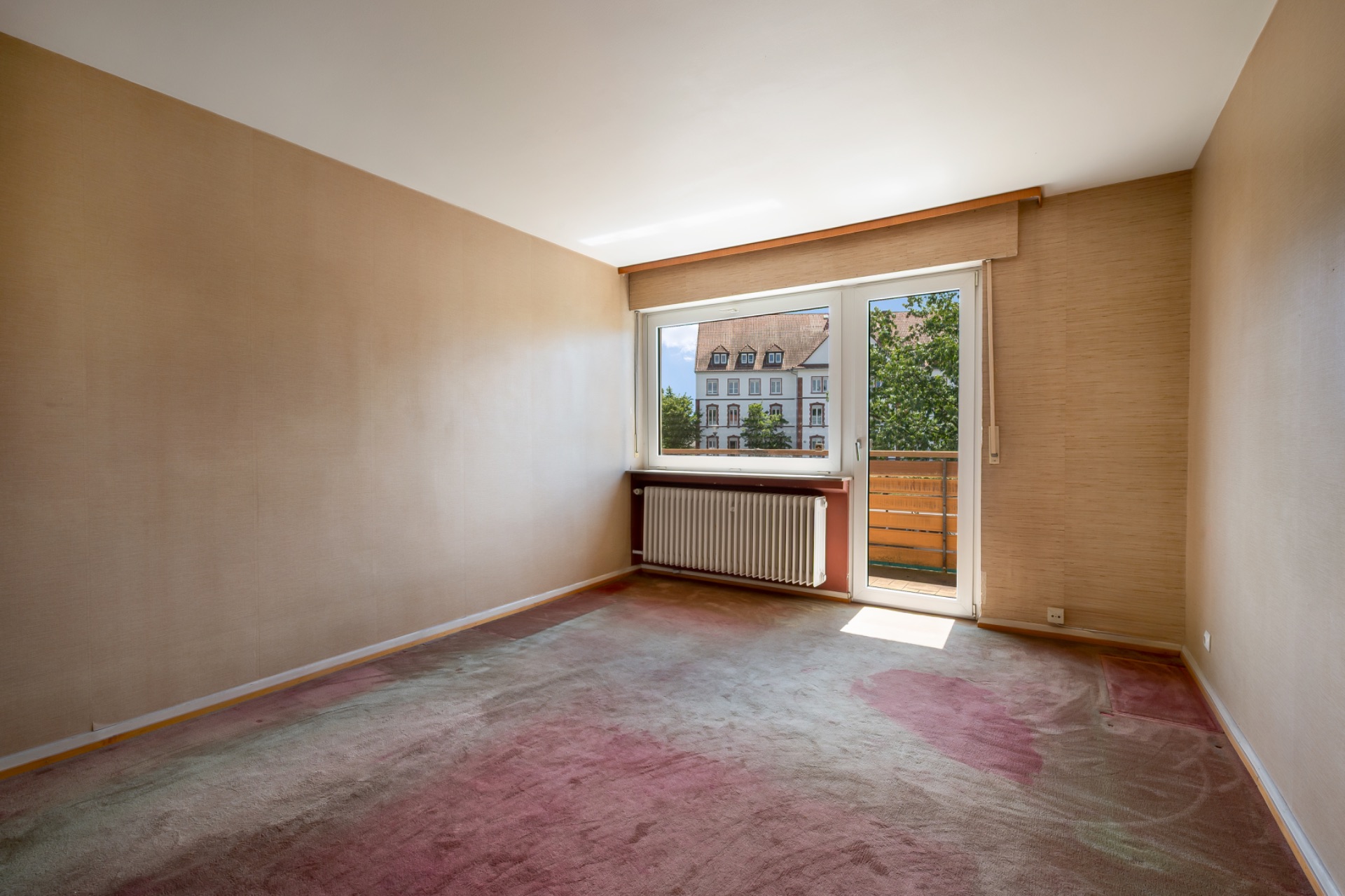 Leeres Zimmer mit Teppichboden, Fenster und Tür zum Balkon, Blick auf Gebäude draußen.
