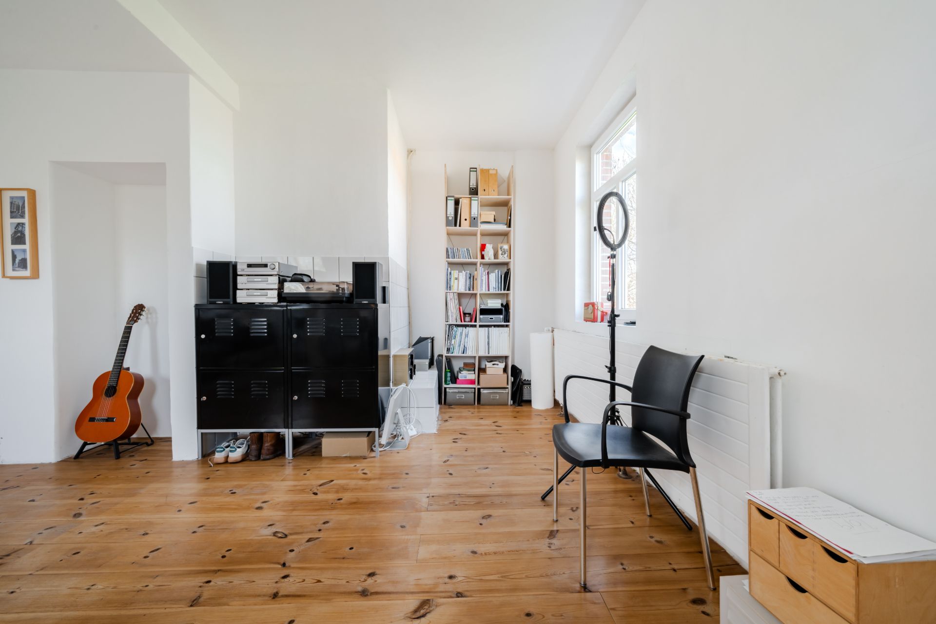 Helles Zimmer mit Holzboden, Regal, Gitarre, schwarzem Stuhl und Ringlicht am Fenster.