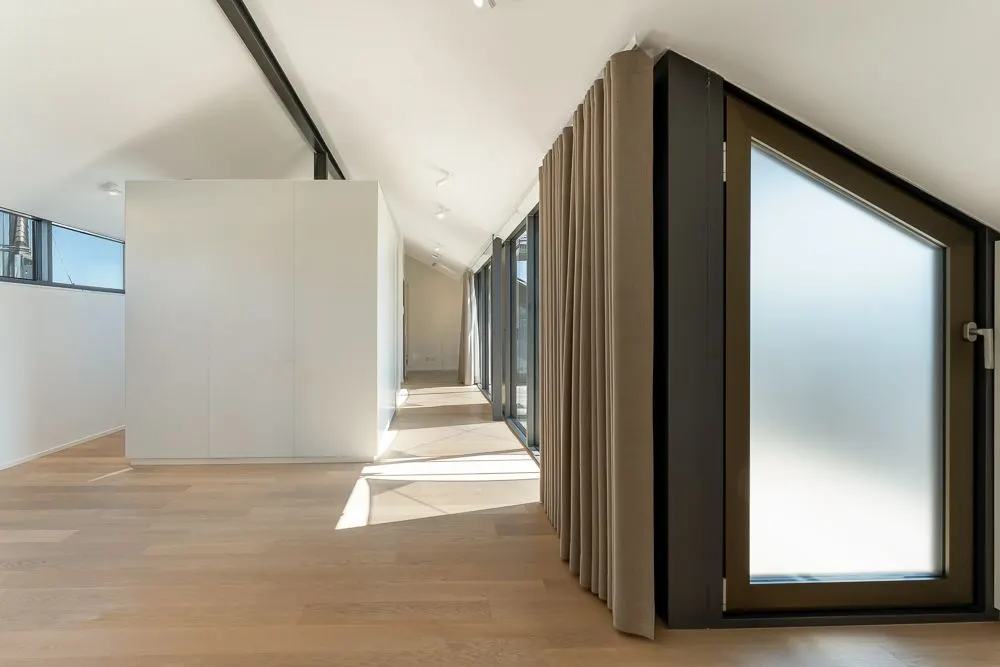 Heller Raum mit schräger Decke, Holzboden, großen Fenstern und Vorhängen.