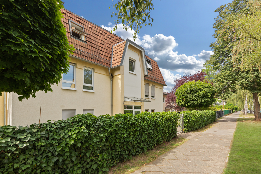 Wohnhaus mit rotem Dach und grüner Hecke an einem Gehweg bei sonnigem Wetter.