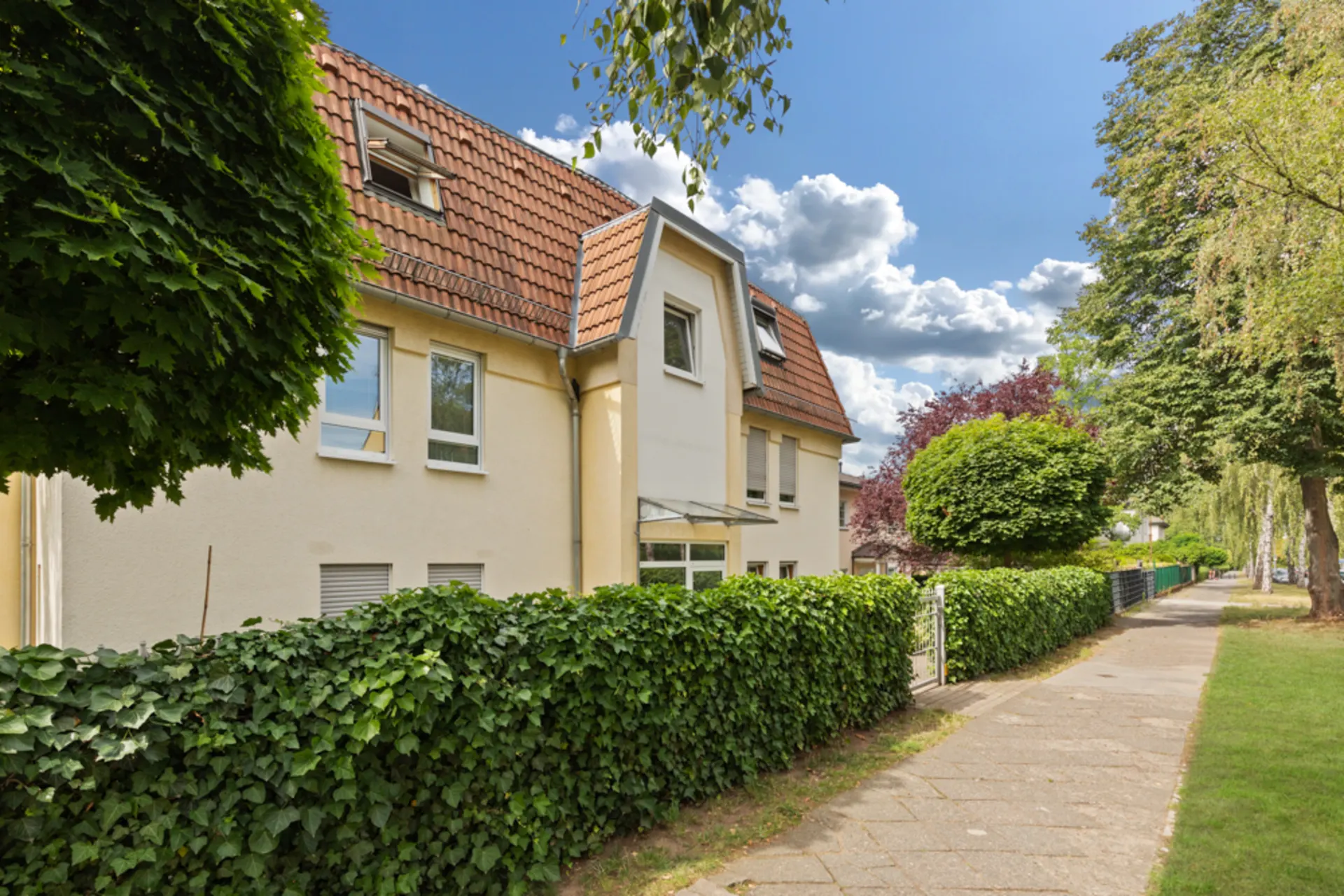 Wohnhaus mit rotem Dach und grüner Hecke an einem Gehweg bei sonnigem Wetter.