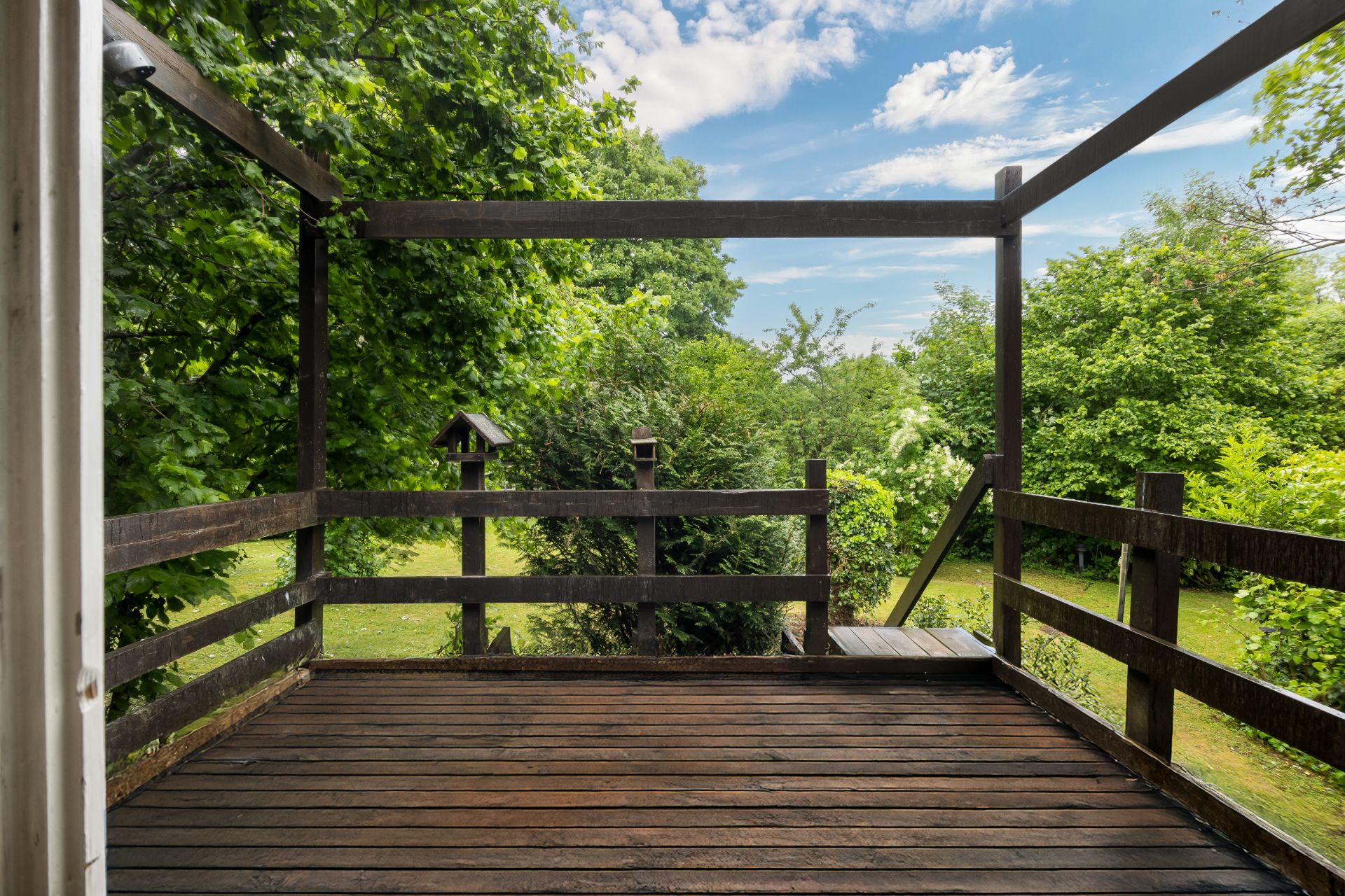 Holzterrasse mit Geländer, Blick auf grünen Garten und Bäume bei blauem Himmel.