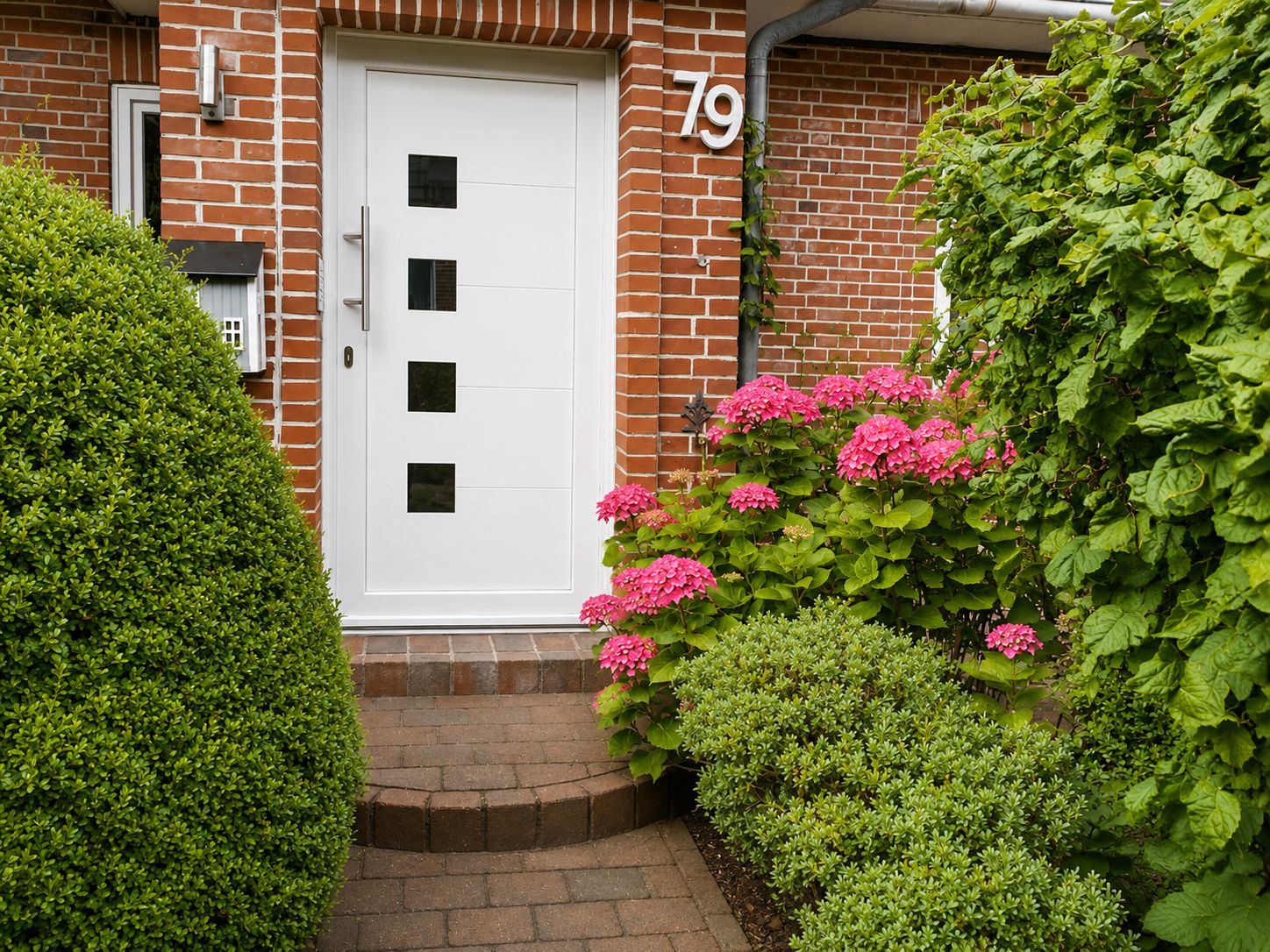 Weiße Haustür mit vier Fenstern, Hausnummer 79, umgeben von grünen Büschen und pinken Blumen.