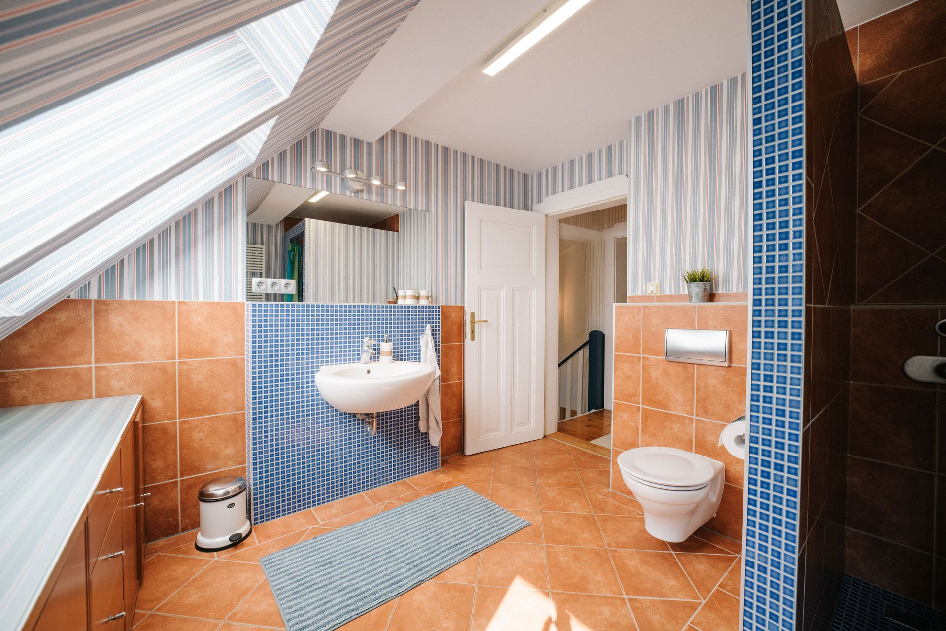 Modernes Badezimmer mit Dachfenster, Waschbecken, WC, Dusche und gestreifter Tapete.