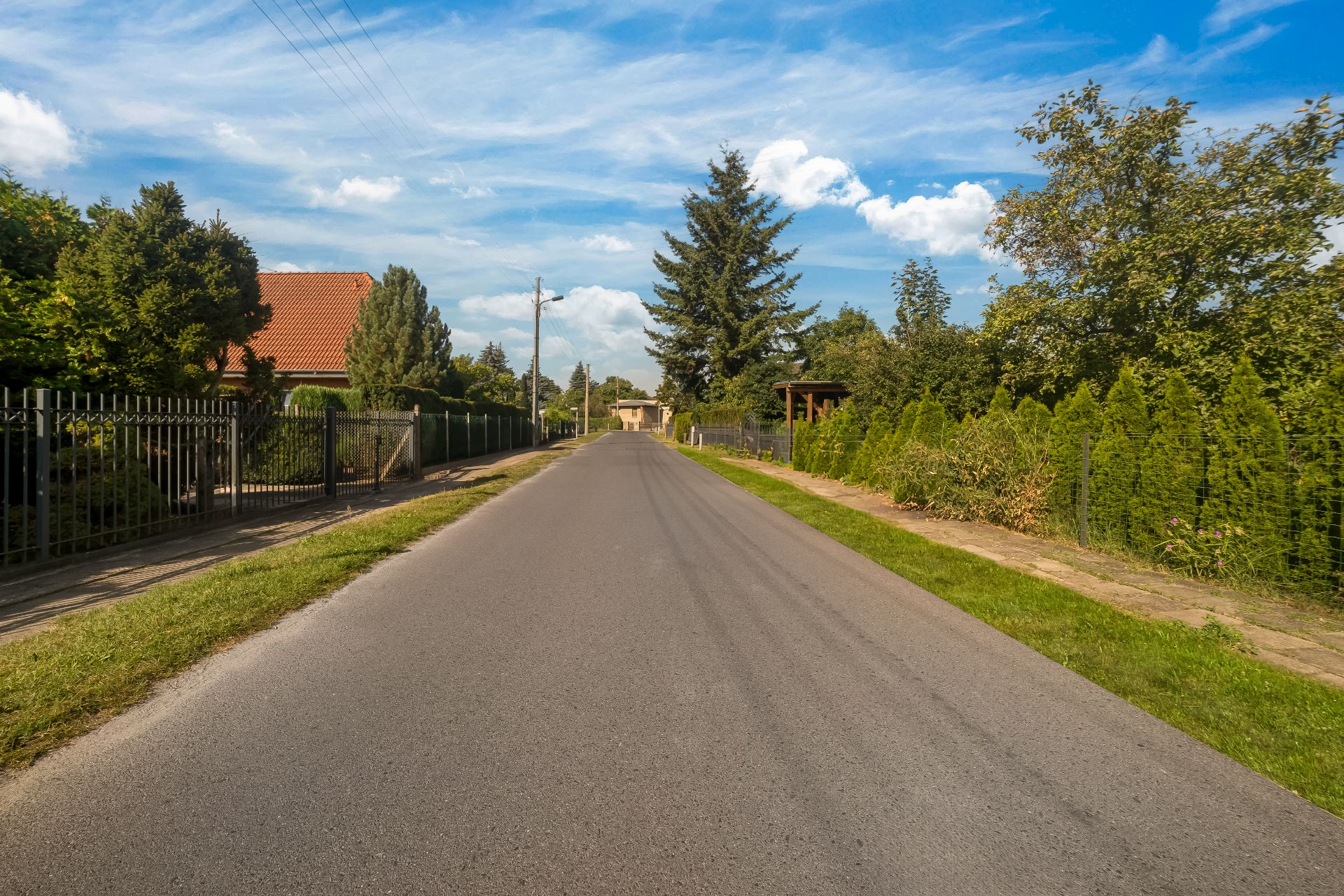 Asphaltierte Straße mit Zäunen und Gärten, gesäumt von Bäumen unter blauem Himmel.