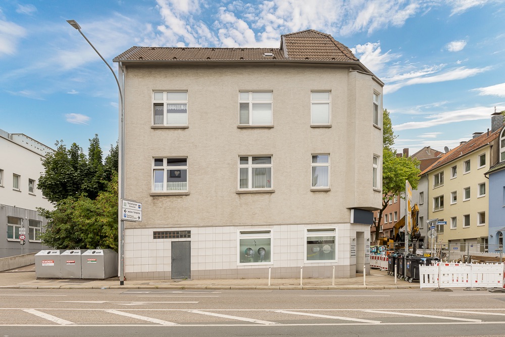 Dreistöckiges Wohnhaus mit beigen Wänden, mehreren Fenstern und braunem Dach an einer Straßenecke.