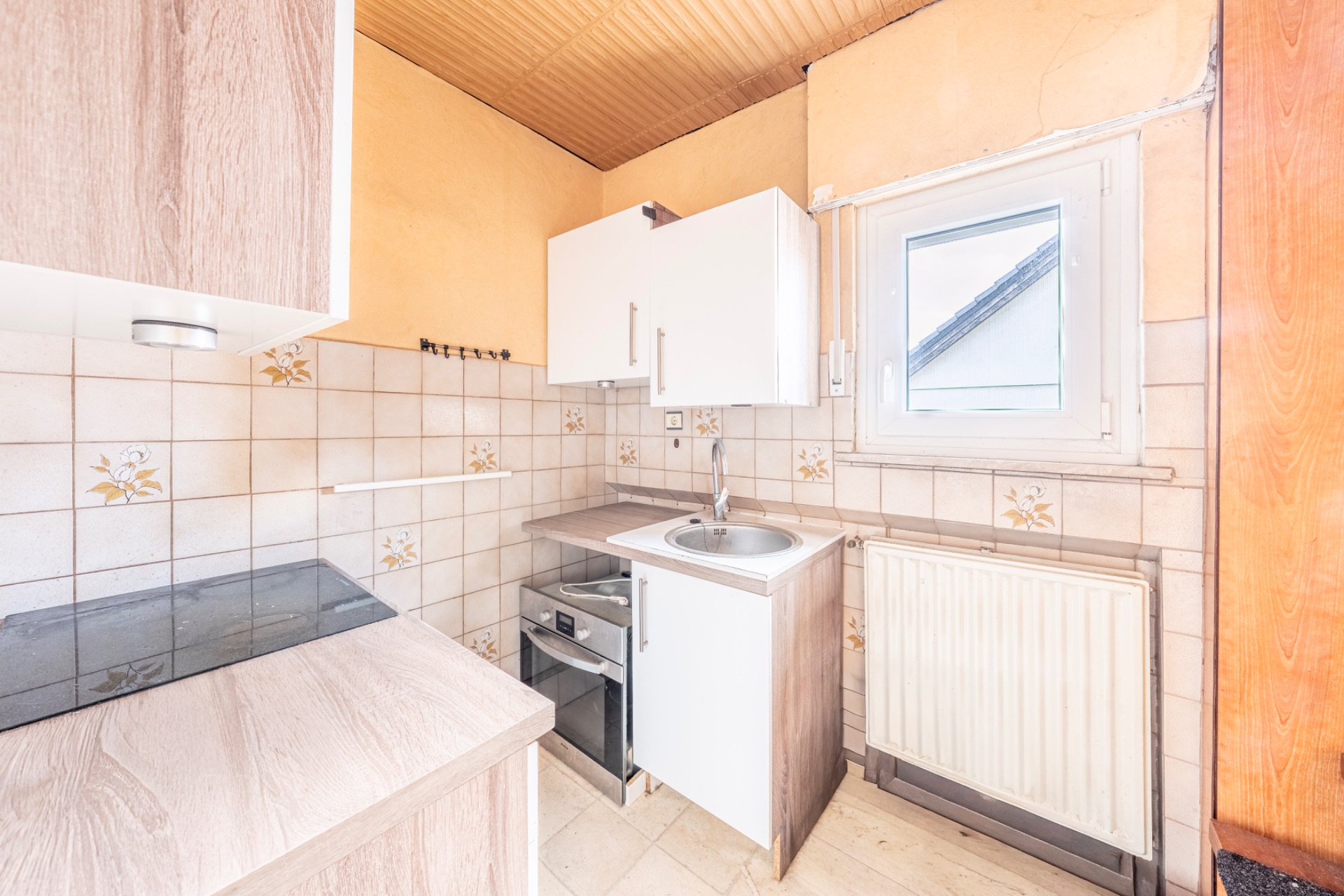 Kleine Küche mit Herd, Spüle, Heizung, Ober- und Unterschränken sowie einem Fenster.