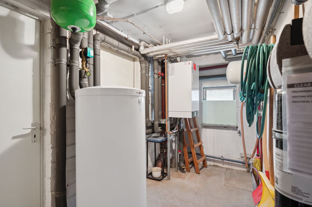 Heizungsraum mit Boiler, Rohren, grüner Ausdehnungsgefäß und Gartenschlauch an der Wand.
