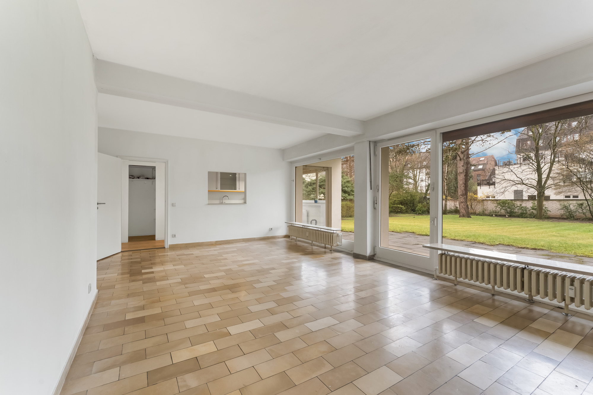 Heller leerer Raum mit großen Fenstern, Fliesenboden und Blick auf Garten.