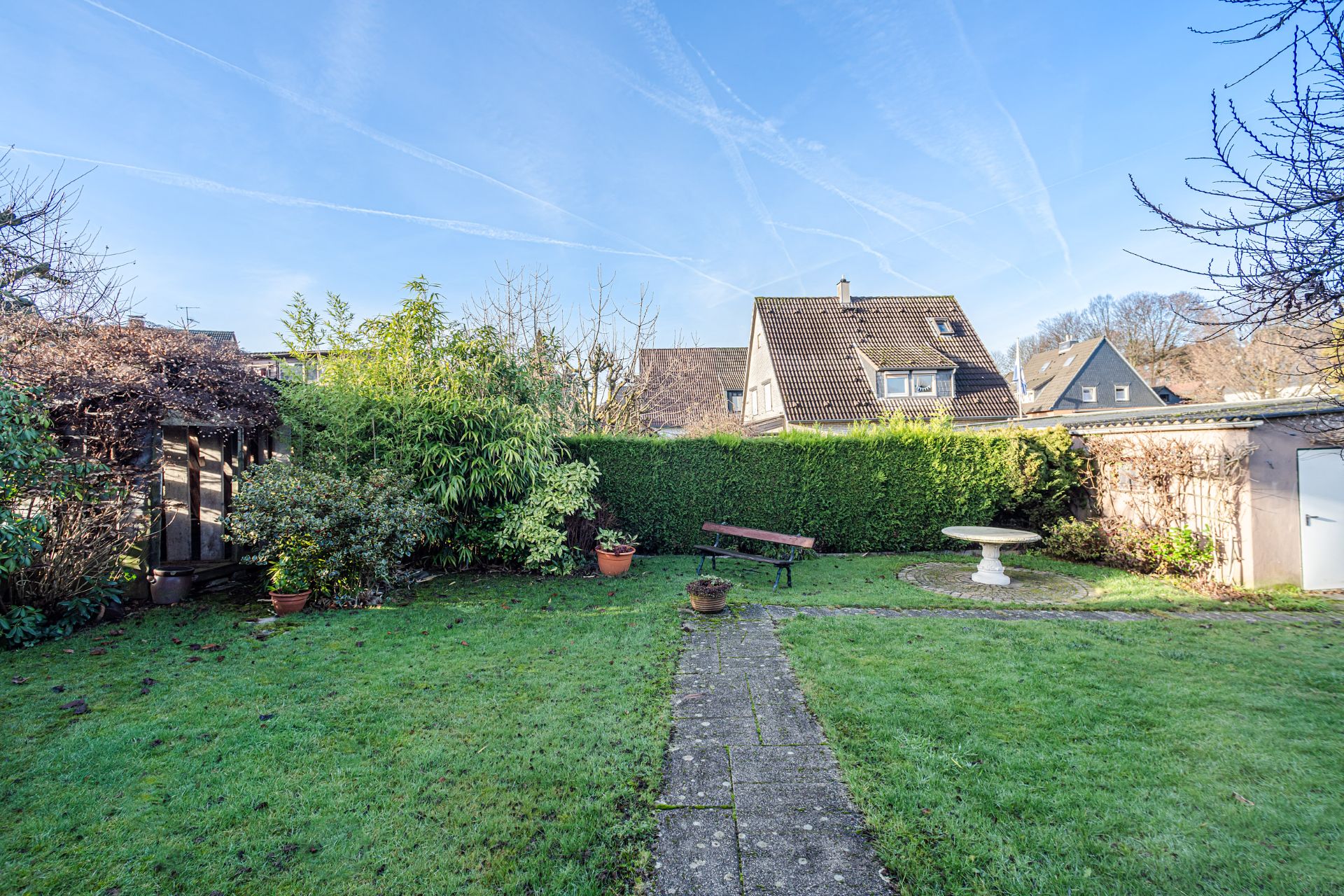 Grüner Garten mit Steinweg, Bank, Vogeltränke und Gartenhaus bei blauem Himmel.