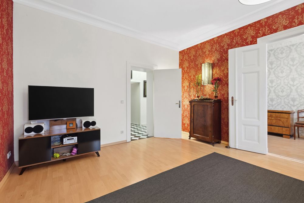 Wohnzimmer mit Fernseher, roter gemusterter Tapete und Holzkommode, Türen zu weiteren Räumen offen.