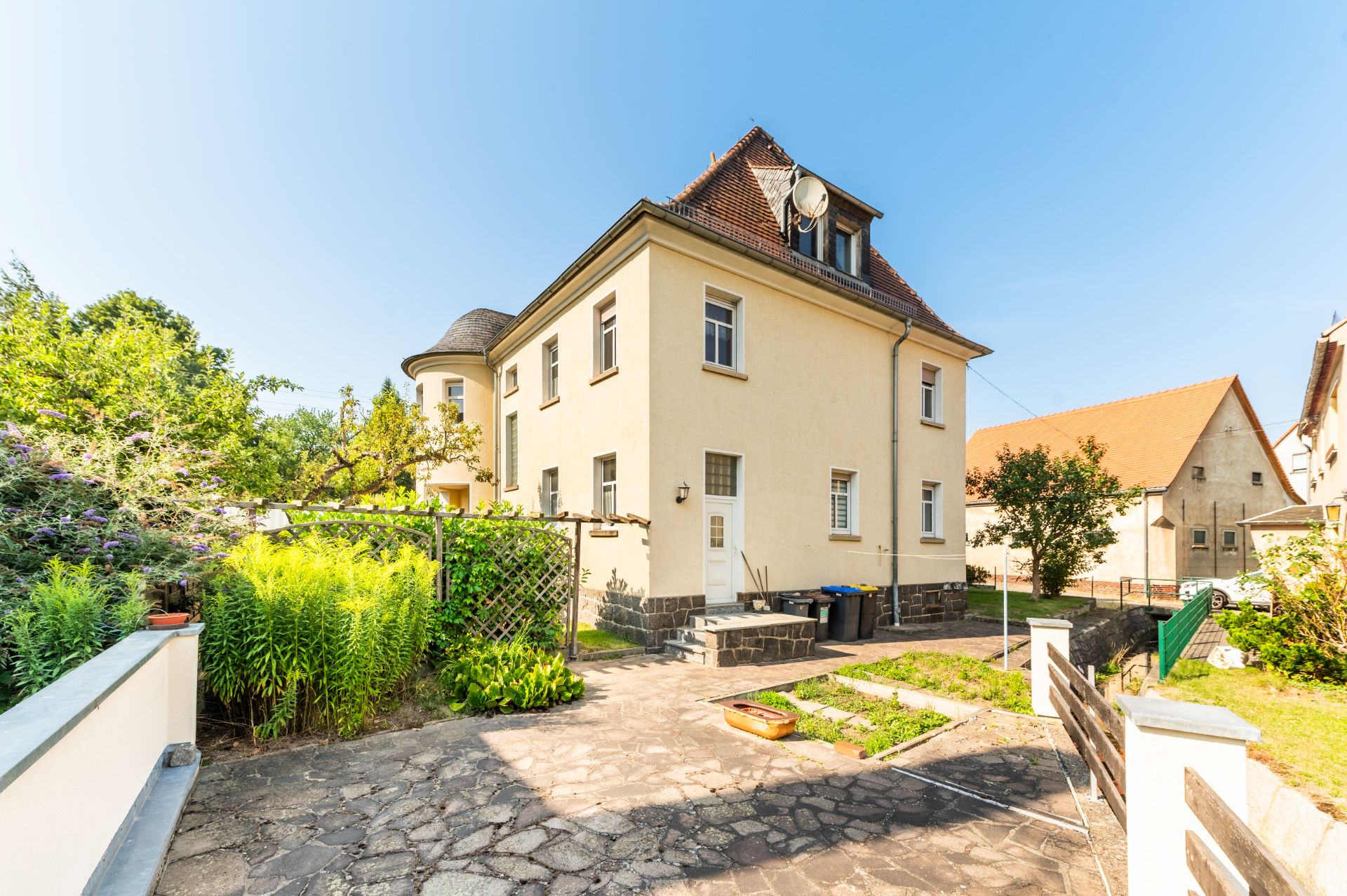 Zweistöckiges Wohnhaus mit Garten, Terrasse und gepflastertem Hof bei sonnigem Wetter.